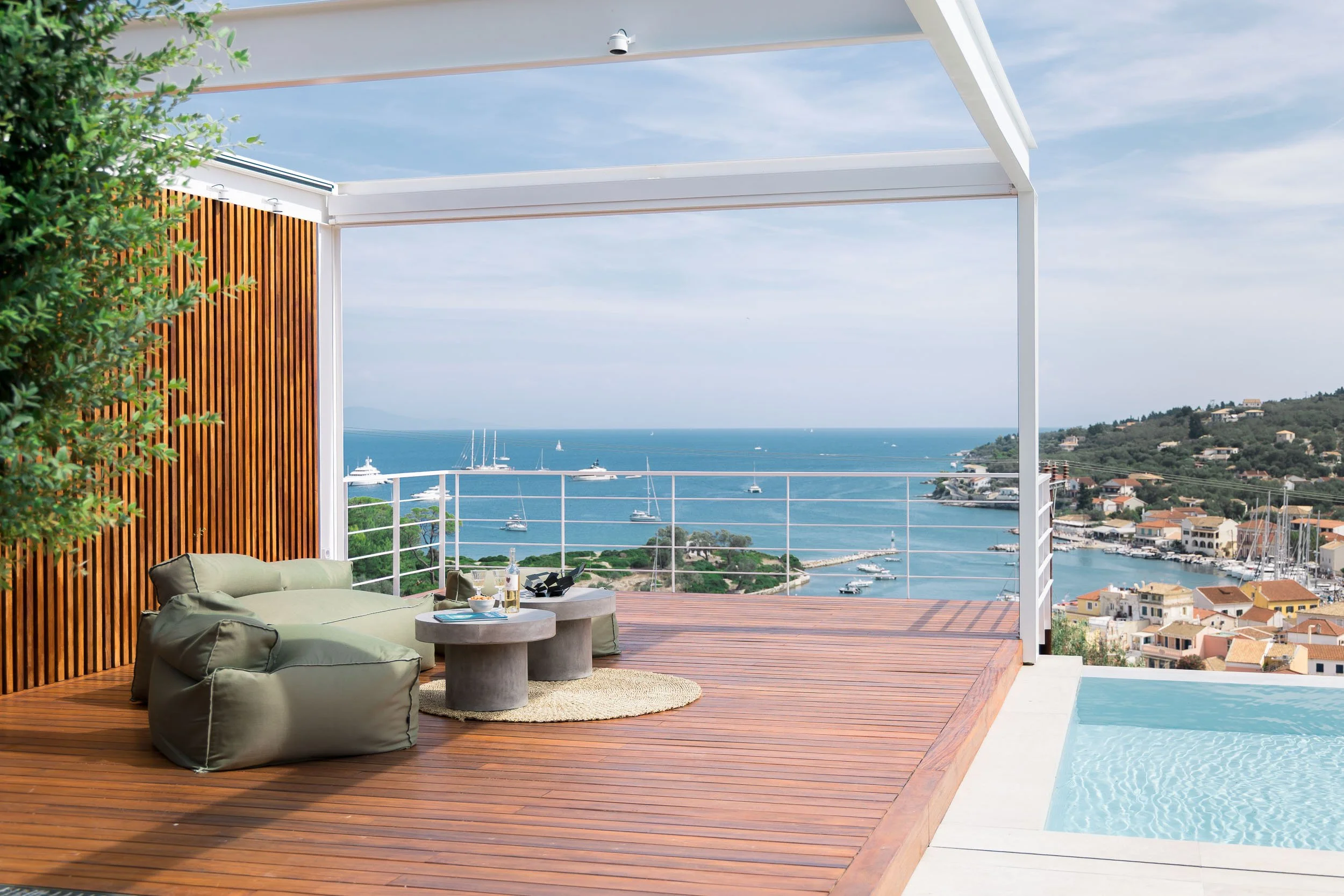 Rooftop balcony with wooden flooring, outdoor seating including bean bags and a small round table, overlooking a marina with boats and yachts on the water, surrounded by hilly terrain and buildings, with a clear blue sky.