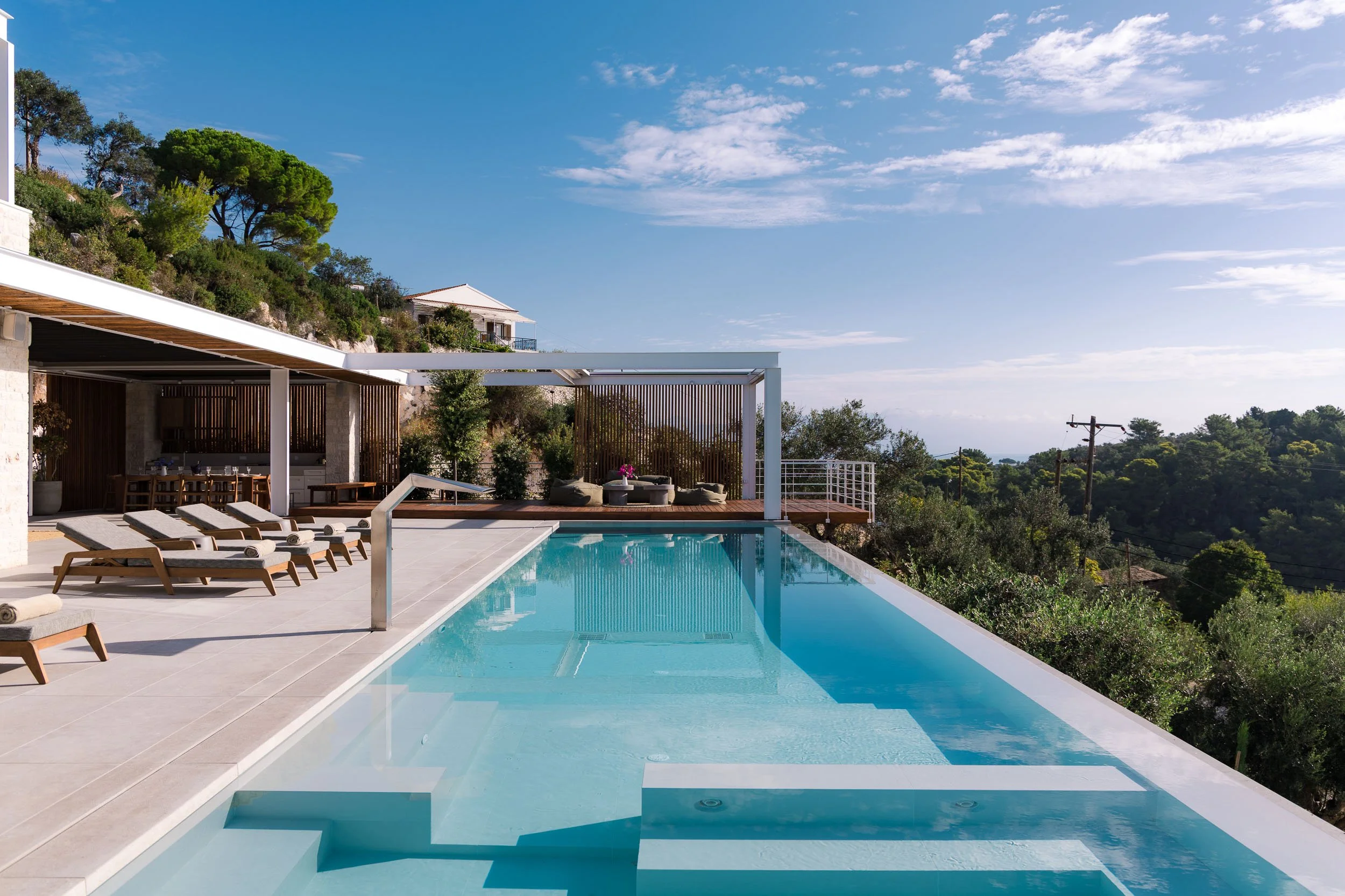 Luxury outdoor hotel pool area with lounge chairs, shaded seating area, surrounding greenery, and distant hills under a partly cloudy sky.