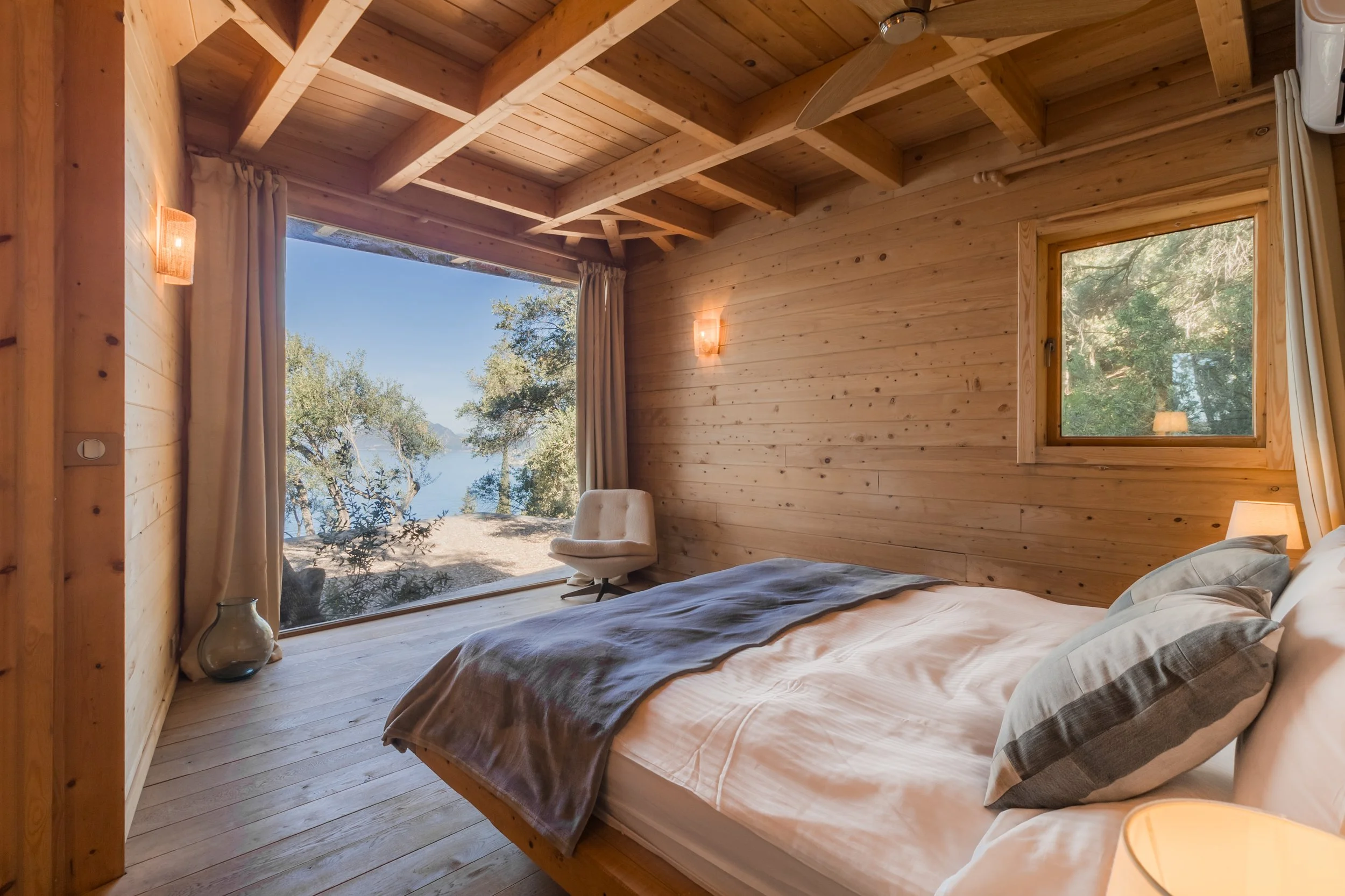 Bedroom with wooden walls and ceiling, large window with a view of trees and water, bed with pillows and blanket, nearby chair and vase.