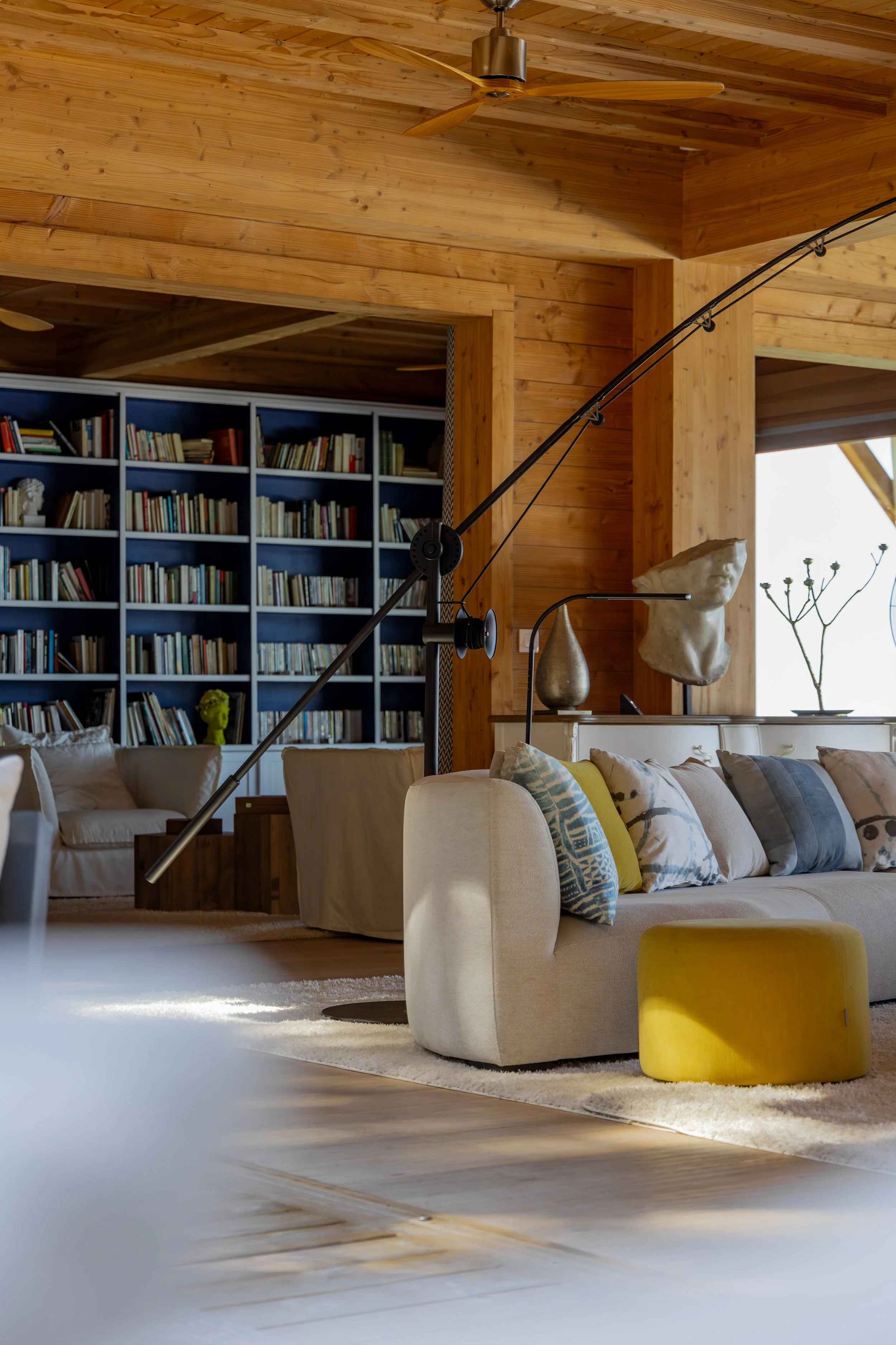 Interior of a living room with wooden walls and ceiling, featuring a beige sofa with colorful pillows, a yellow ottoman, sculptures, and a large bookshelf filled with books.