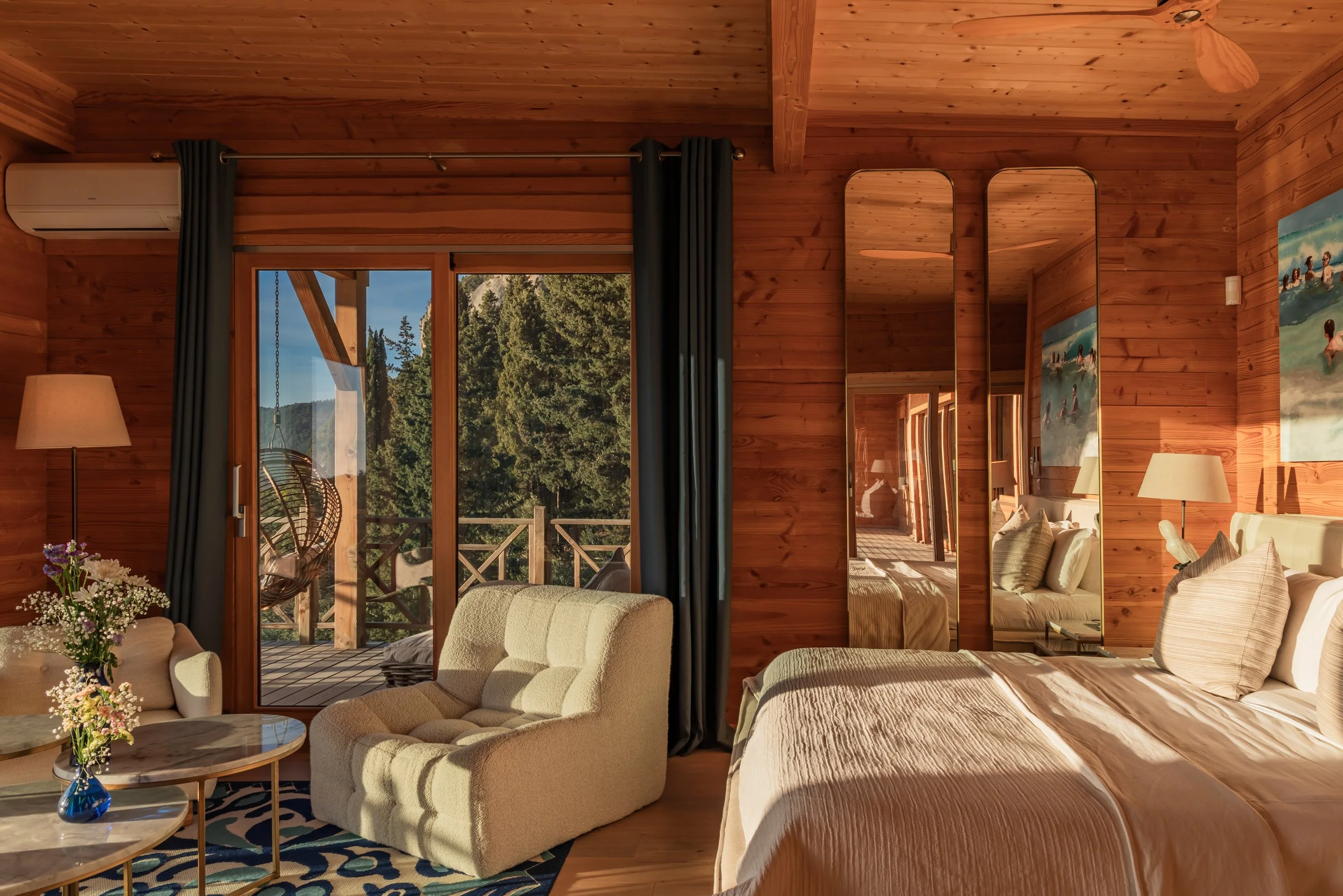 A cozy bedroom with wooden walls and ceiling, featuring a large bed with beige bedding, a nightstand with a lamp, a mirror, and a sliding glass door leading to a balcony with trees outside. There is also a small sitting area with a beige armchair and