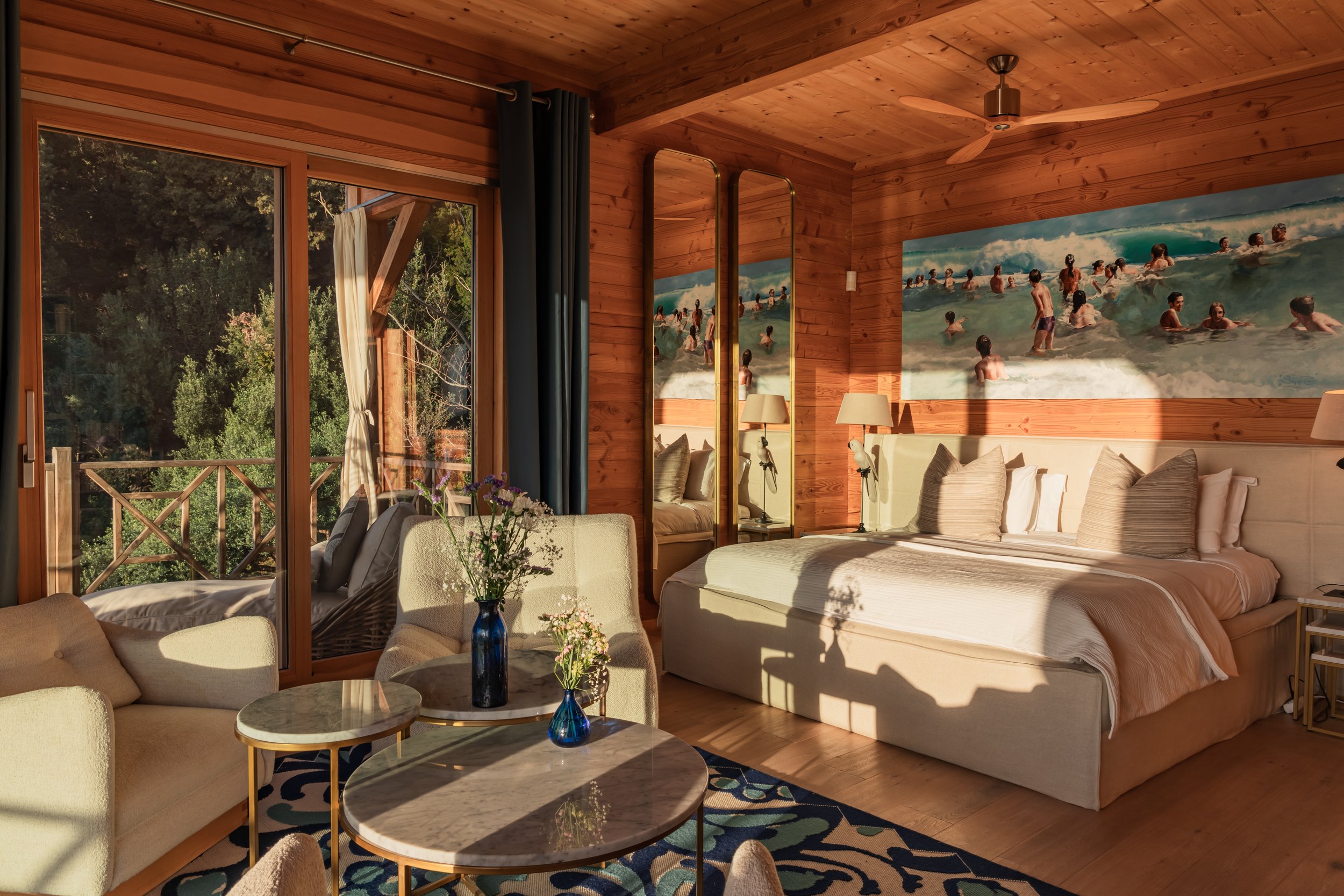 A cozy bedroom with wooden walls and ceiling, featuring a large bed with cream-colored linens, two mirrors, and wall art of people at the beach. There's a sitting area with a sofa and round coffee tables near a large sliding glass door leading to a b