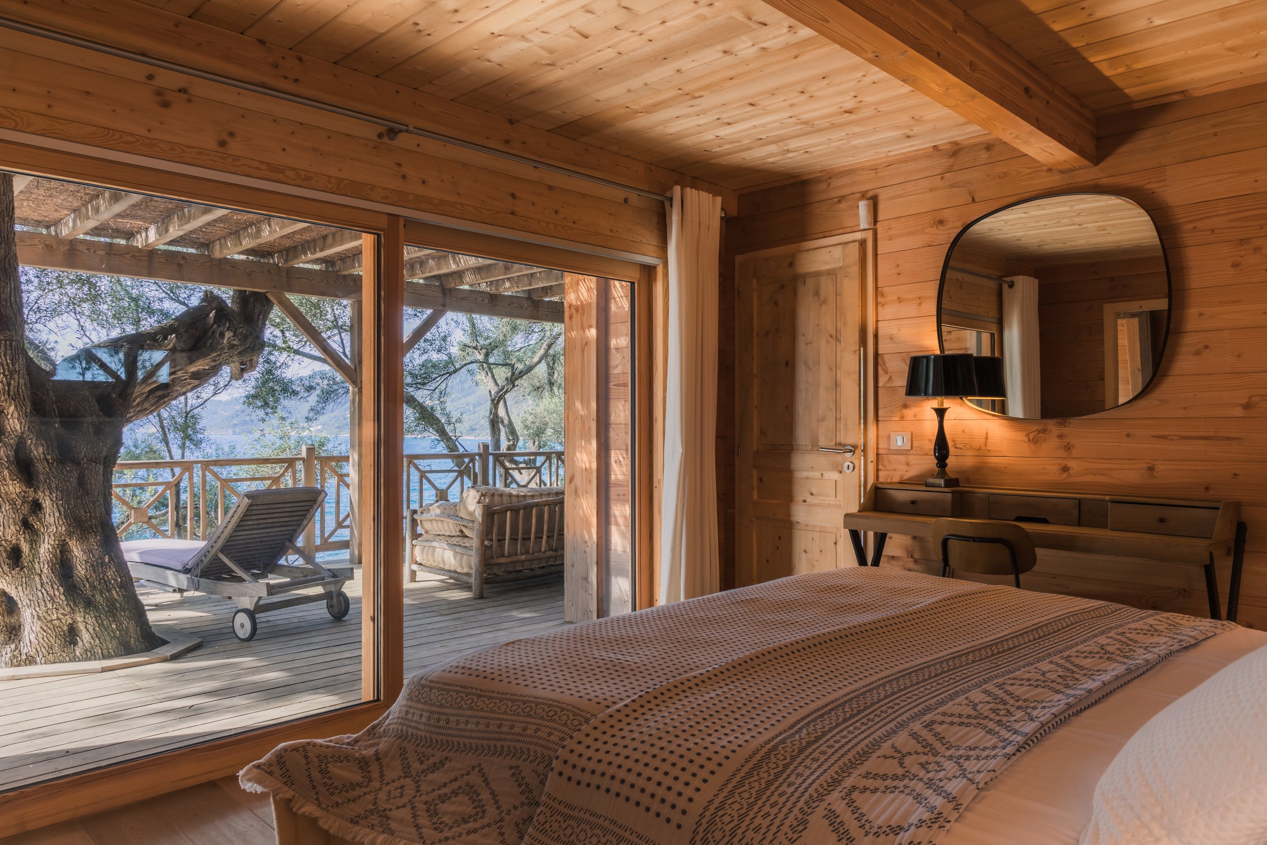 A cozy bedroom with wooden walls and ceiling, a bed with patterned blanket, a round mirror on the wall, a black lamp, and a desk with a chair. Outside, there is a wooden deck with outdoor furniture, a lounge chair, a sofa, surrounding trees, and a vi