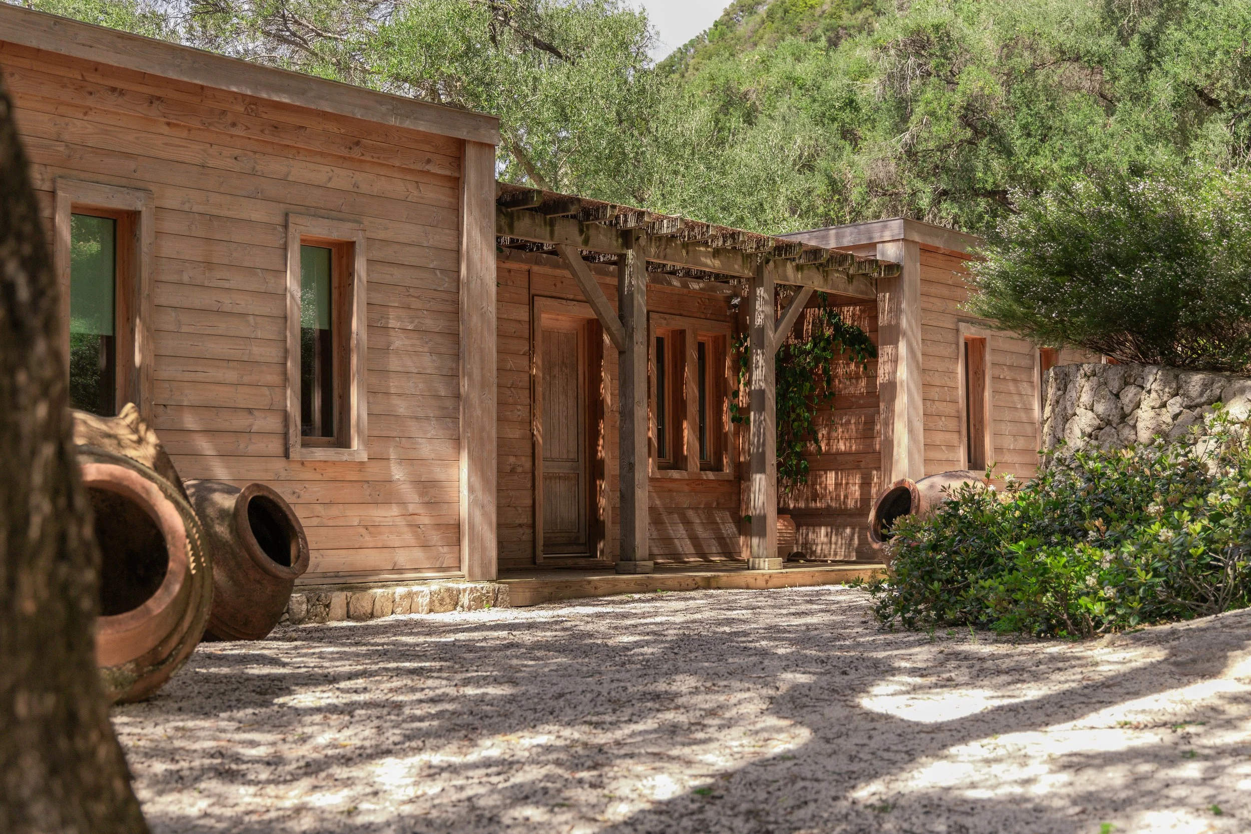 A rustic wooden cabin surrounded by greenery and trees, with empty clay pots on the gravel ground outside.