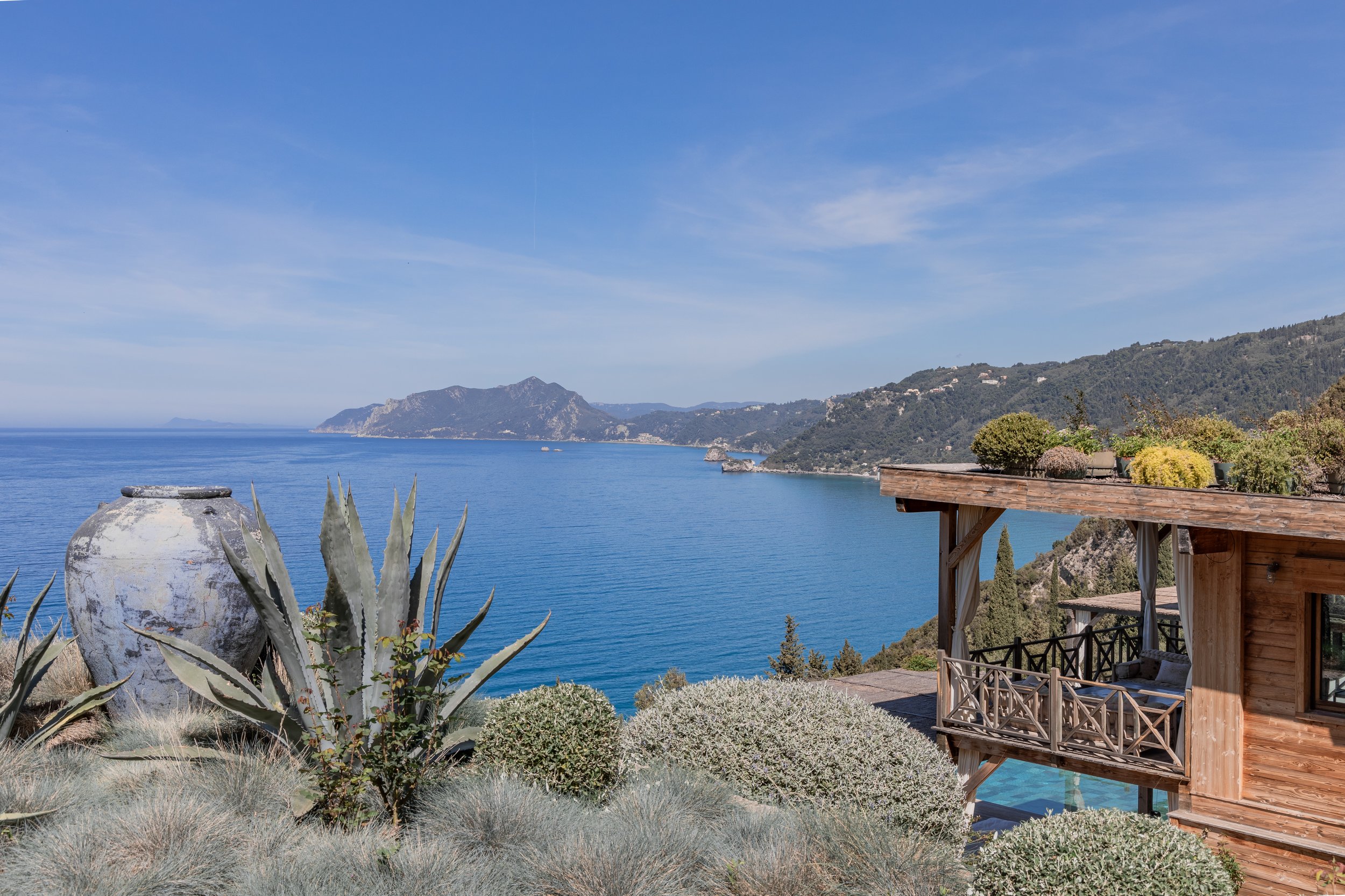 Seaside view of a coastal landscape with a wooden balcony and garden plants, overlooking calm blue water and distant mountains under a clear sky.