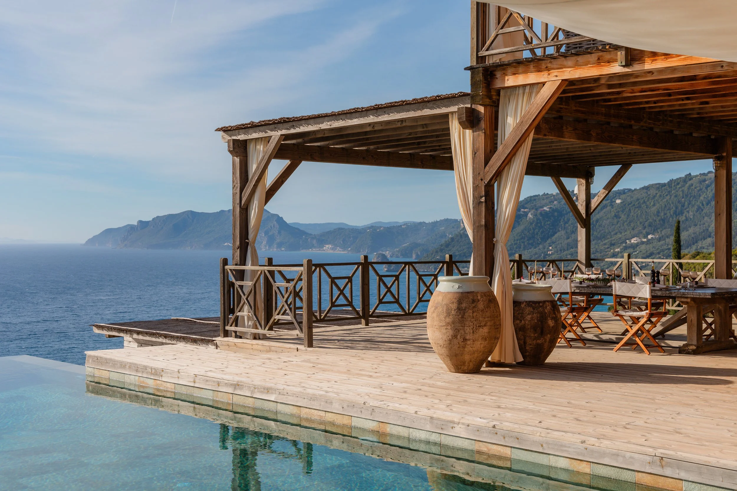 A wooden deck with a dining area, overlooking an ocean with mountains in the background, featuring a pool in the foreground.