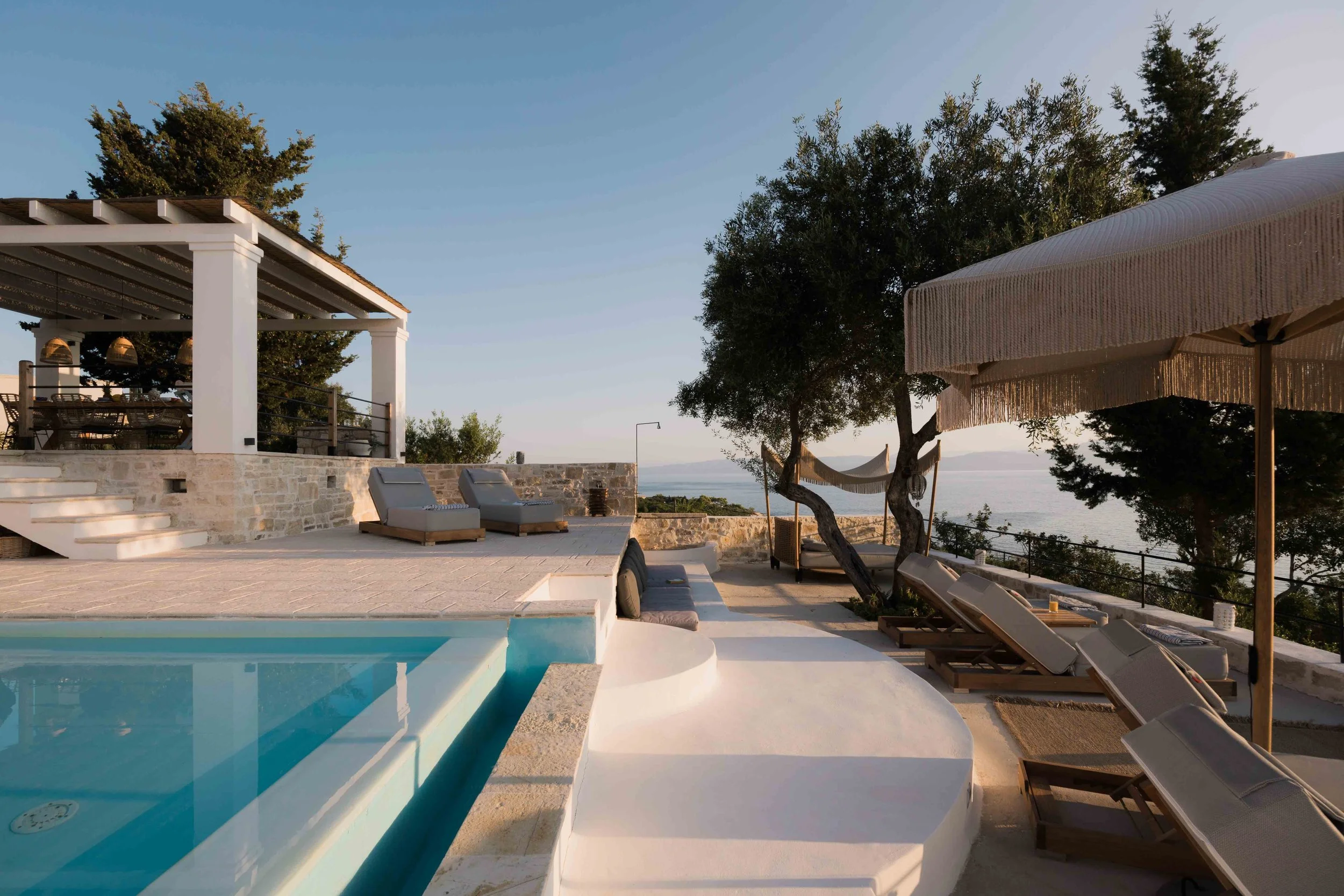 Luxury outdoor pool area with poolside lounge chairs, a shaded dining area, and trees overlooking the ocean at sunset.