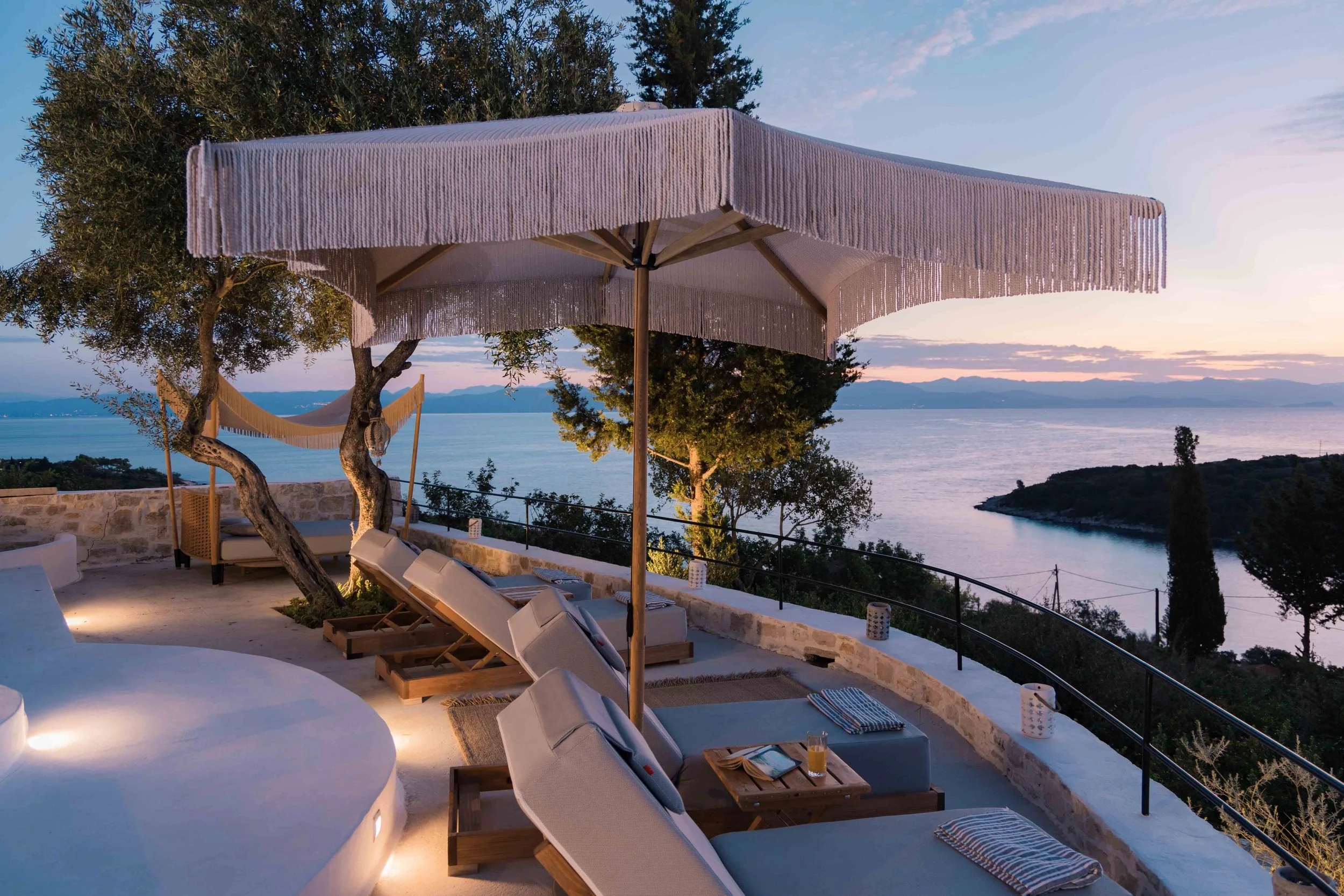 A serene outdoor seating area overlooking the water at sunset with lounge chairs, an umbrella, trees, and a view of islands and mountains in the distance.
