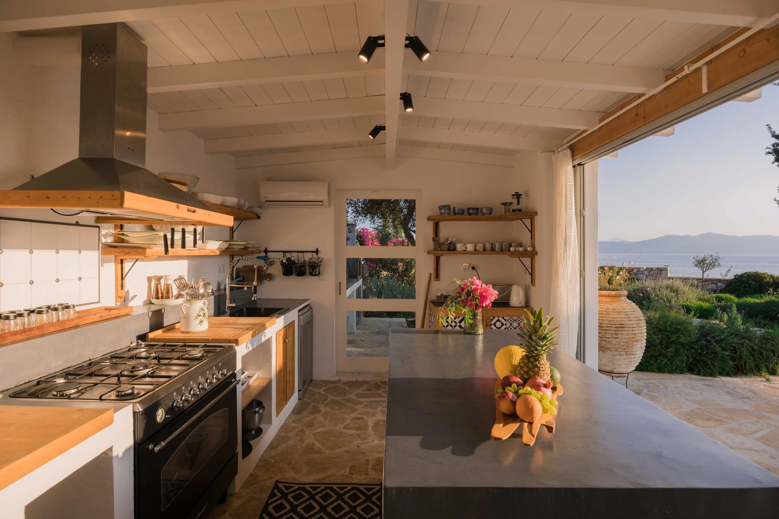 Kitchen with white walls and ceiling, wooden shelves holding dishes and cups, a stove, a sink, a countertop with a pineapple, fruit, and flowers, open sliding door revealing a view of green landscape and mountains by the water.