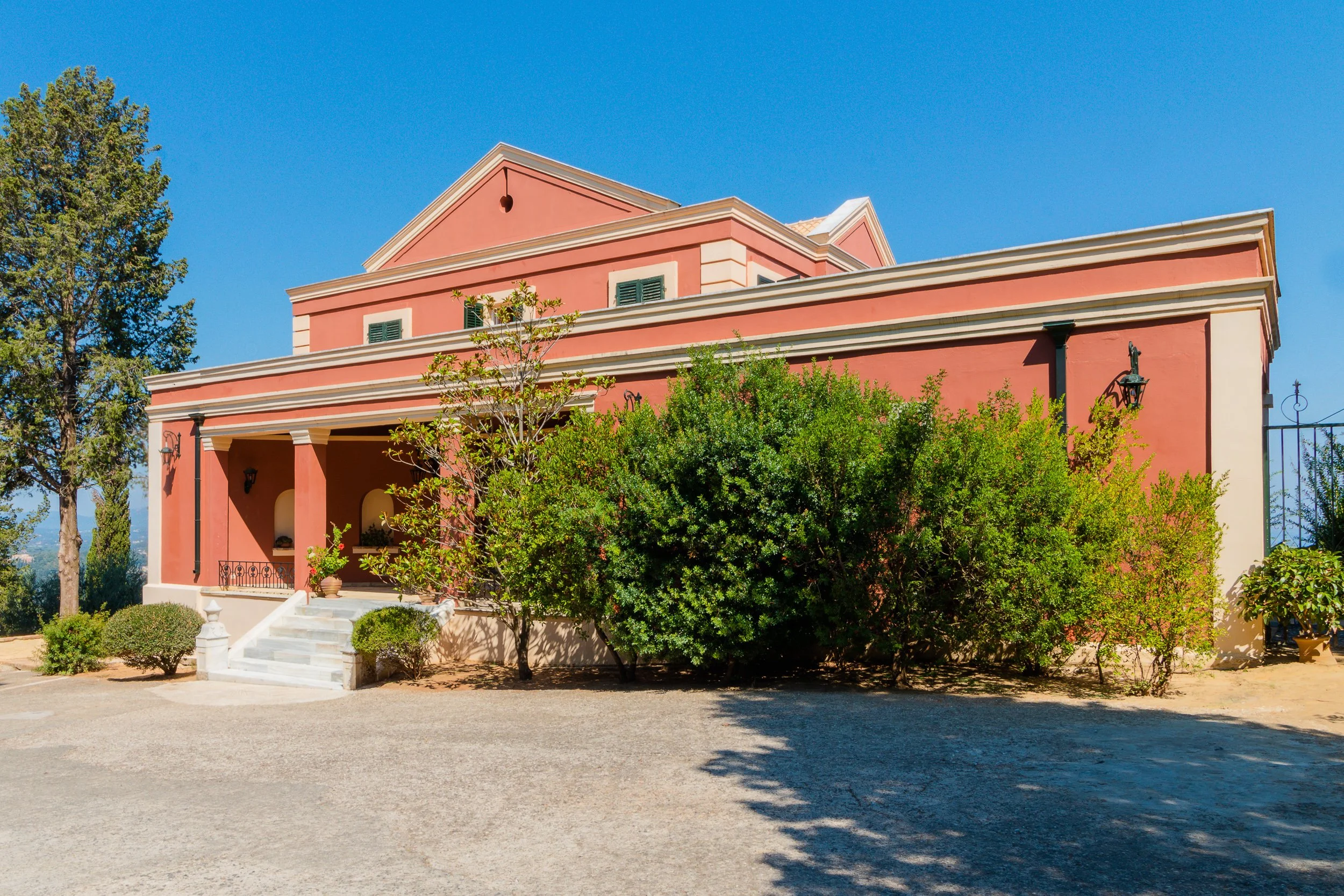 A pink Mediterranean-style house with white accents, a small staircase leading to a porch, surrounded by green shrubs and trees, under a clear blue sky.