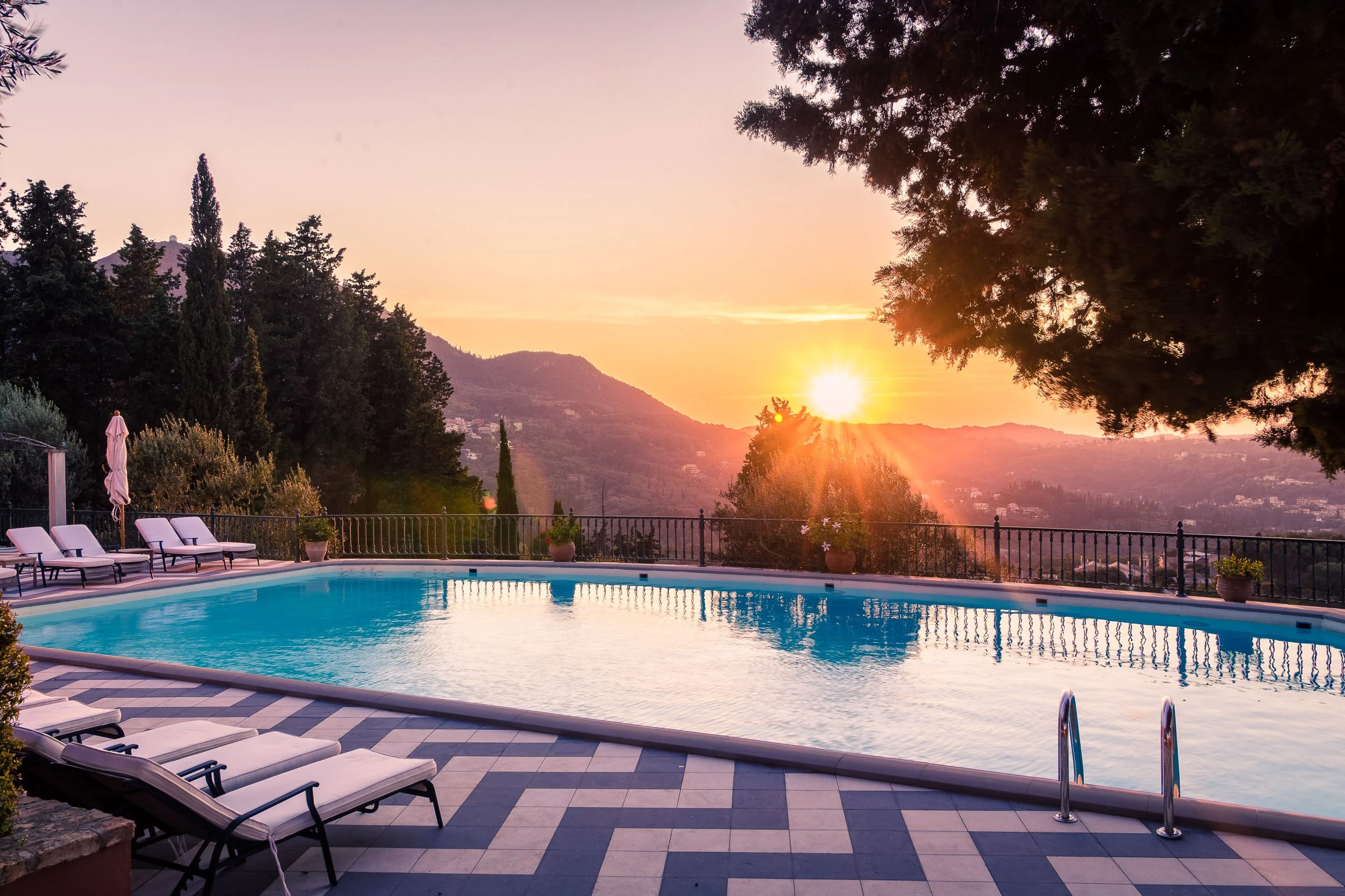 A swimming pool at sunset with lounge chairs and an umbrella on the deck, surrounded by trees and hills in the background.