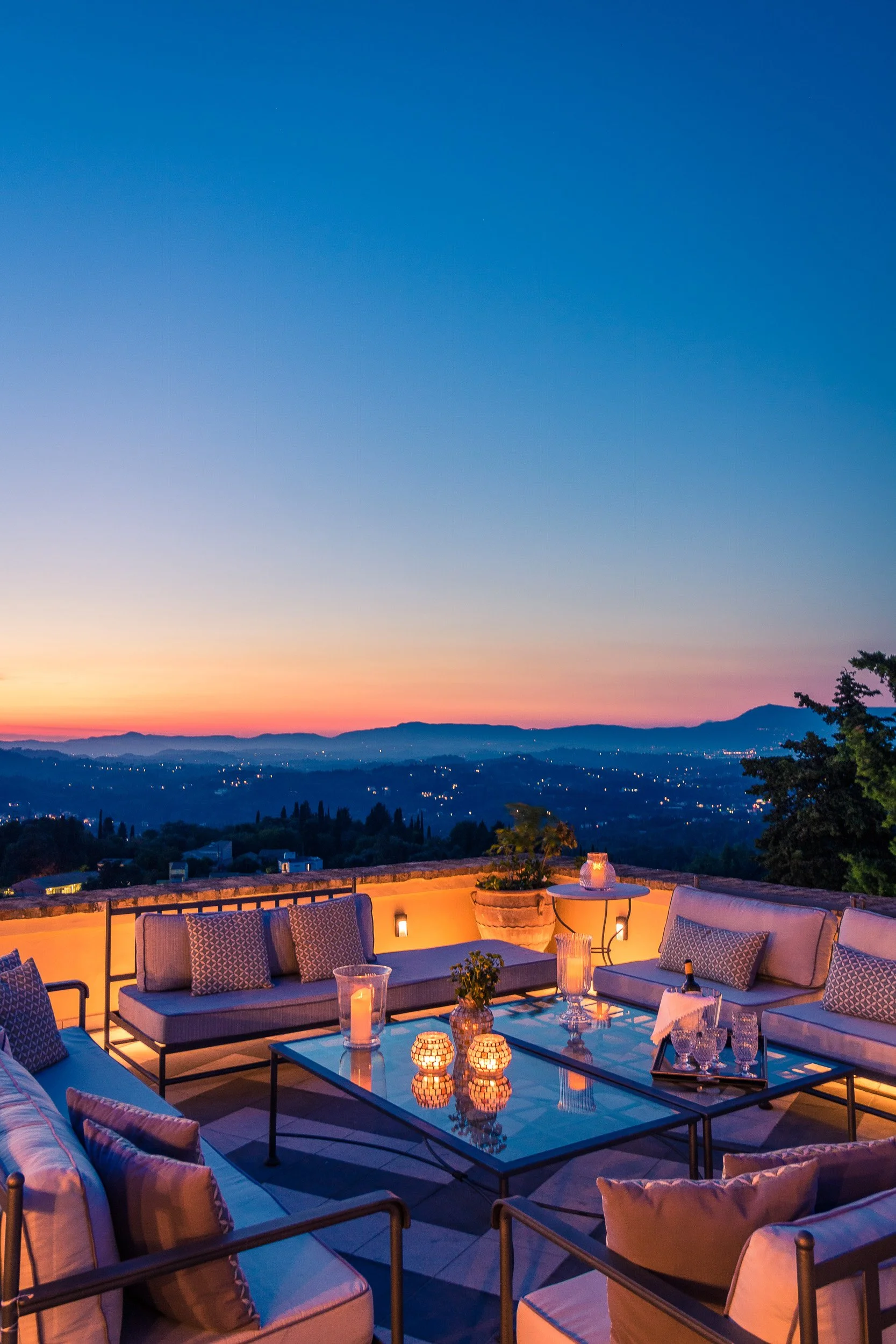 Outdoor patio with cushioned seating and candlelit table overlooking a sunset or twilight view of a valley and mountains in the distance.