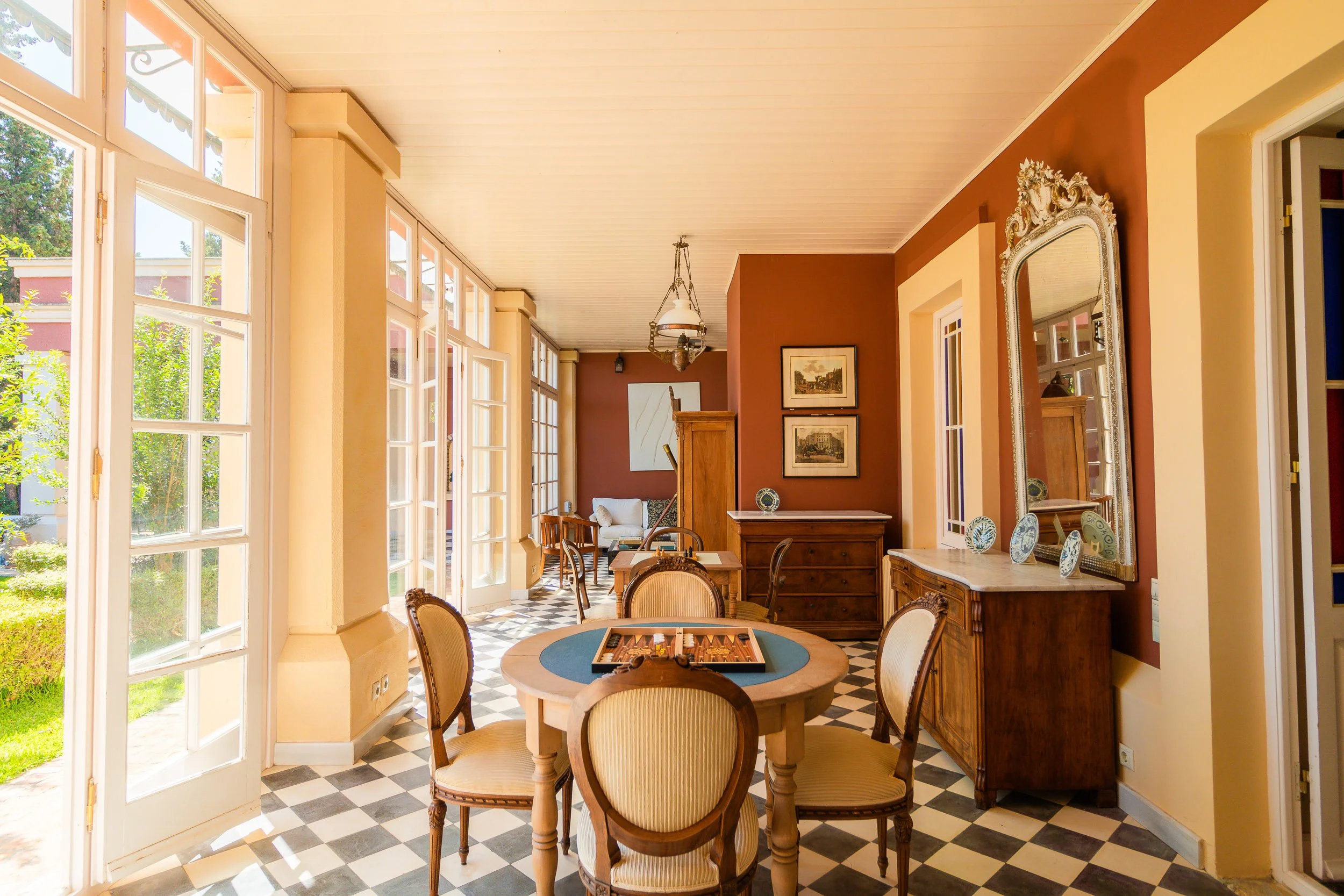 Bright, sunny dining area with checkered black and white tile floor, wooden dining table with six chairs, built-in wooden cabinetry, framed artwork, a large ornate mirror, and a view of green garden outside through glass-paneled doors.