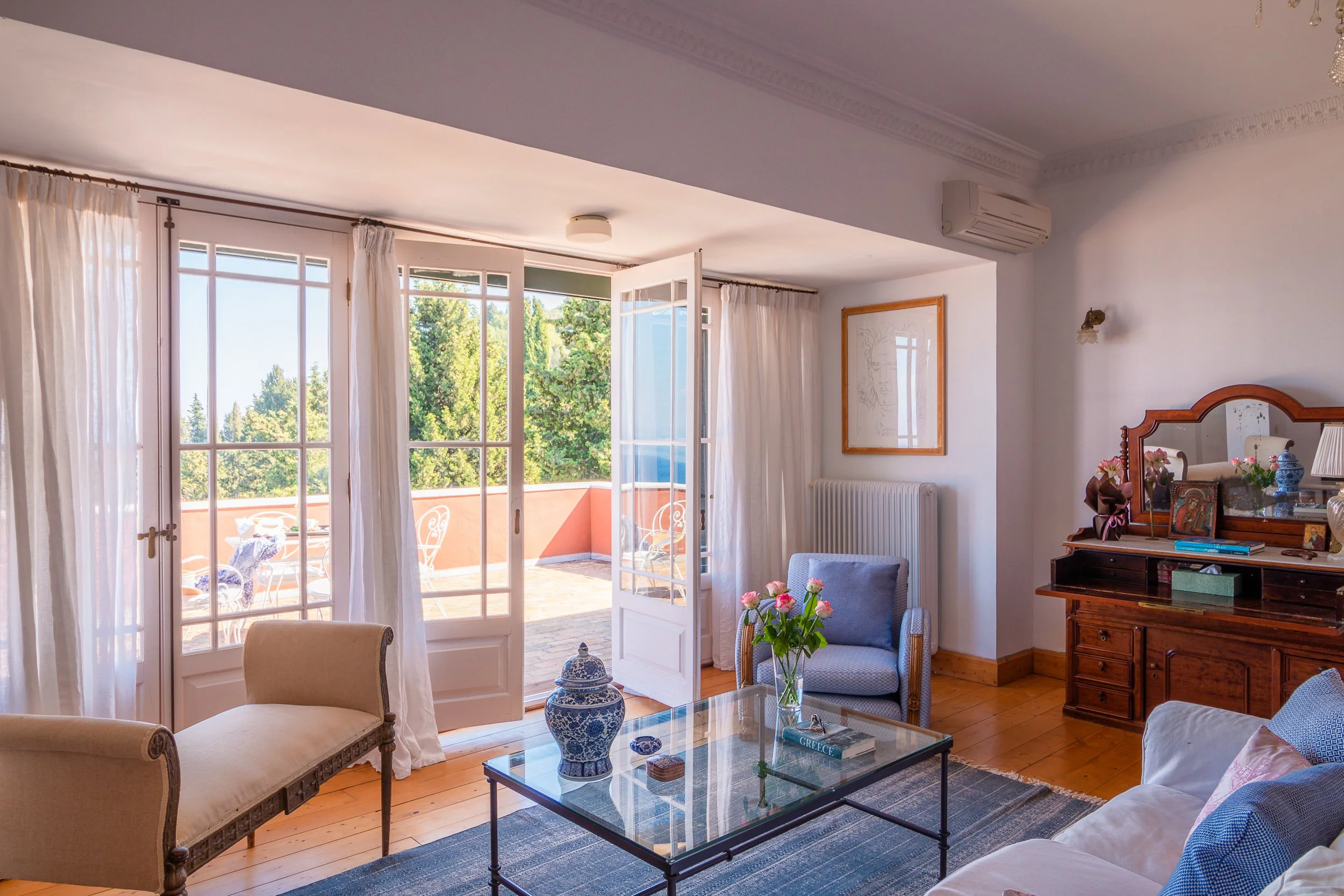 Living room with glass doors leading to balcony, chairs, a glass coffee table with decorative objects, and a wooden dresser with mirror, flowers, and books.