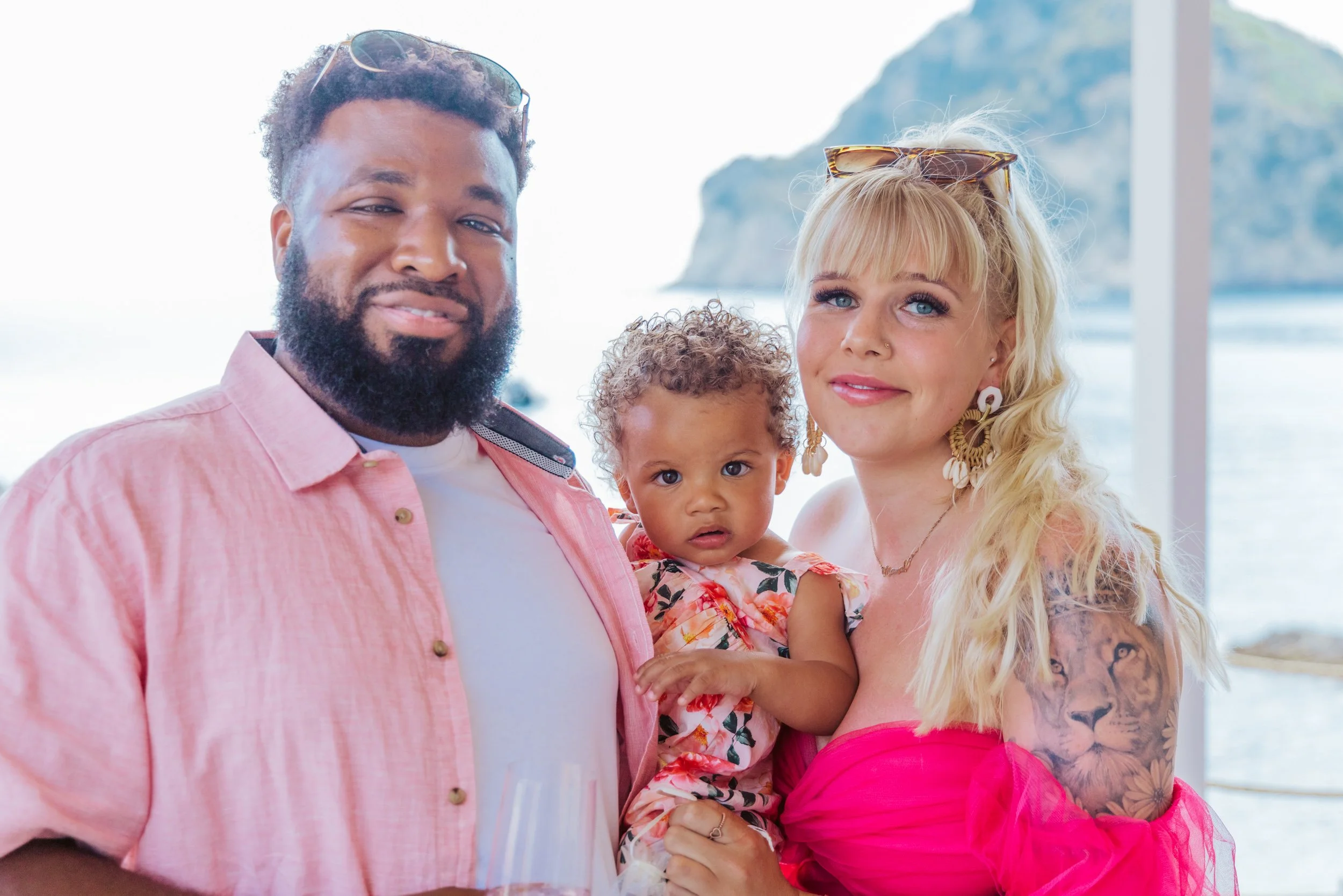 Three people, an African American man with glasses on his head, a blonde woman with tattoos wearing earrings, and a young girl with curly hair, standing together at a beachside location with ocean and cliffs in the background.