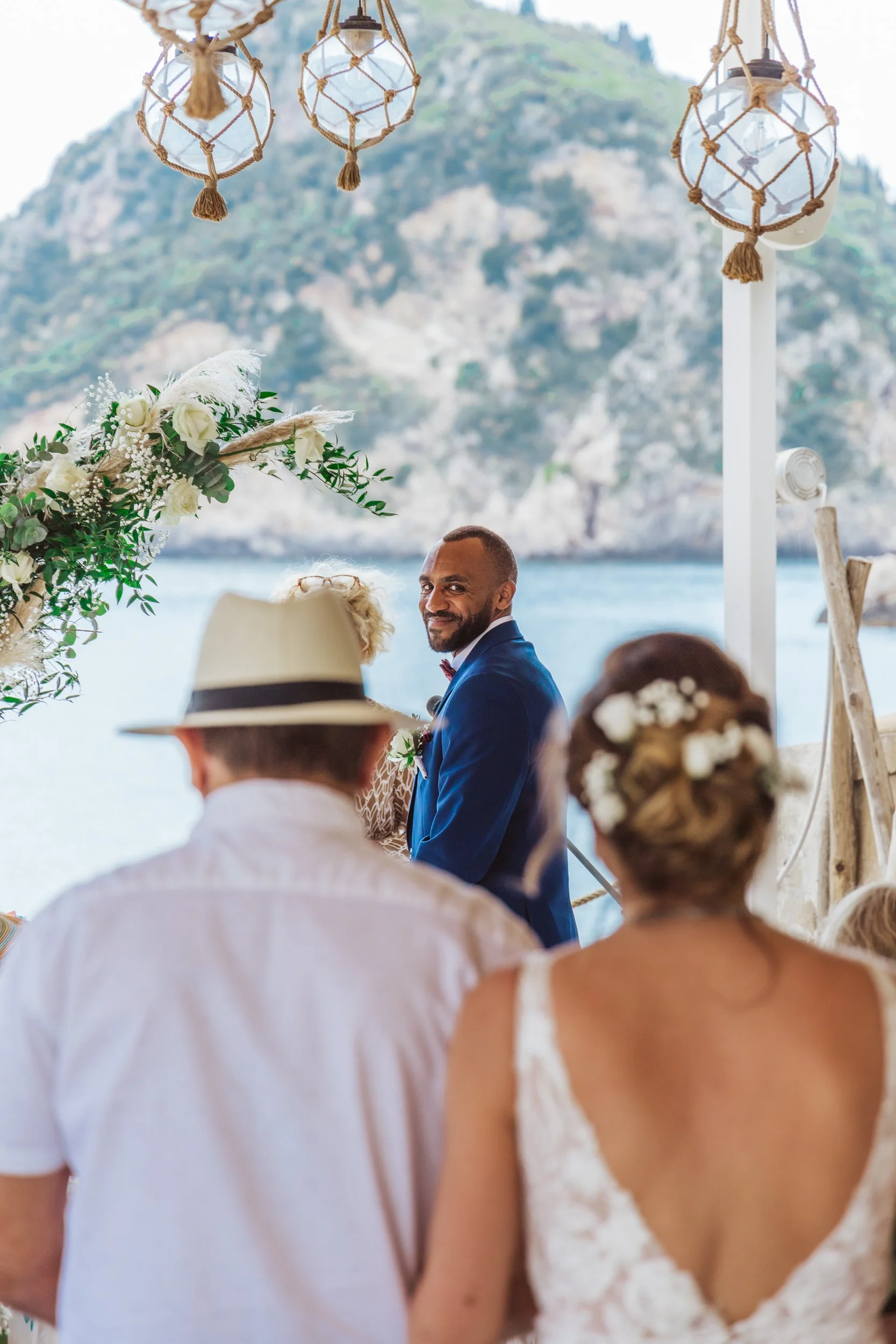 A wedding ceremony taking place outdoors by the water with scenic mountains in the background. The groom, in a blue suit, is smiling at the bride, who has floral hair accessories. Guests are seated, including a man wearing a hat and a woman with flow