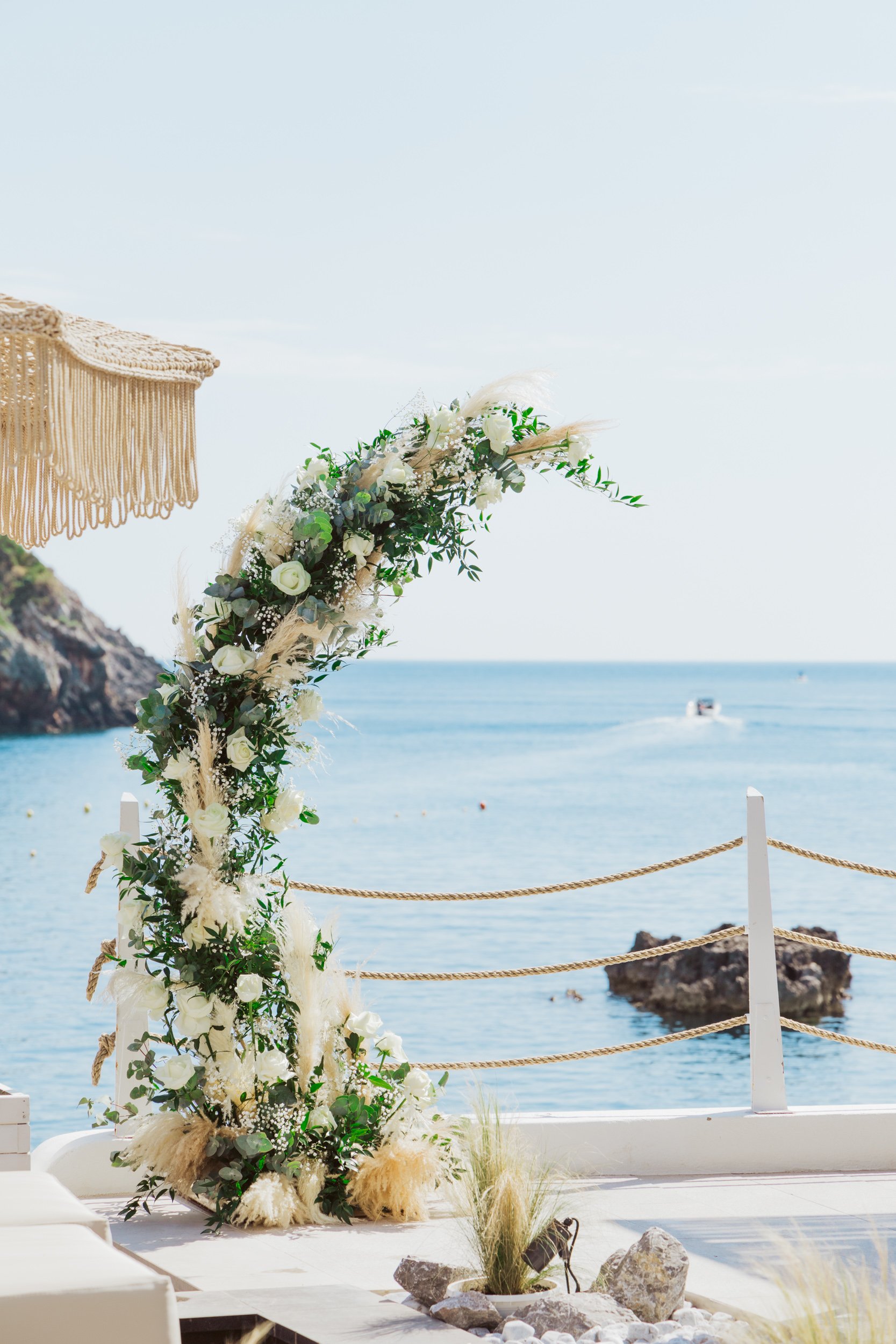 A floral wedding arch with white roses, greenery, and pampas grass overlooking the ocean with a boat in the distance.