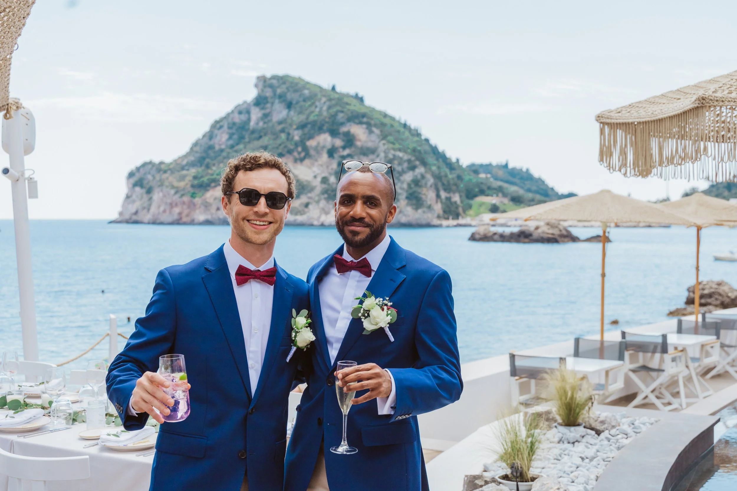 Two men dressed in blue suits with red bow ties and white shirts, wearing boutonnieres, holding drinks, smiling at the camera on a seaside patio with umbrellas and an island in the background.