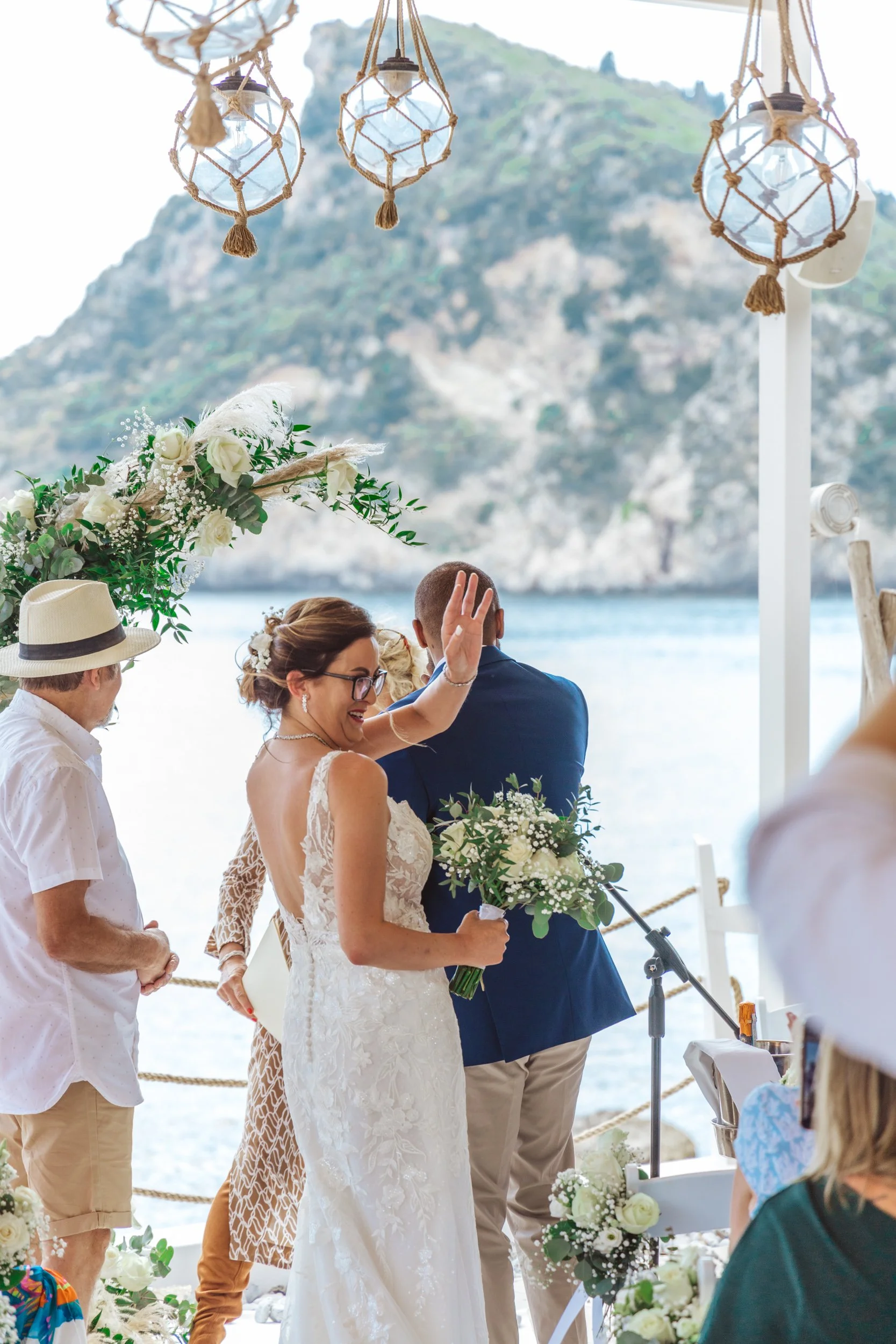 A bride in a white lace wedding dress celebrating at her wedding ceremony by the water, holding a bouquet of white flowers with greenery, while the groom, in a navy blue suit, faces away. Guests and decorative elements like hanging lanterns and flora