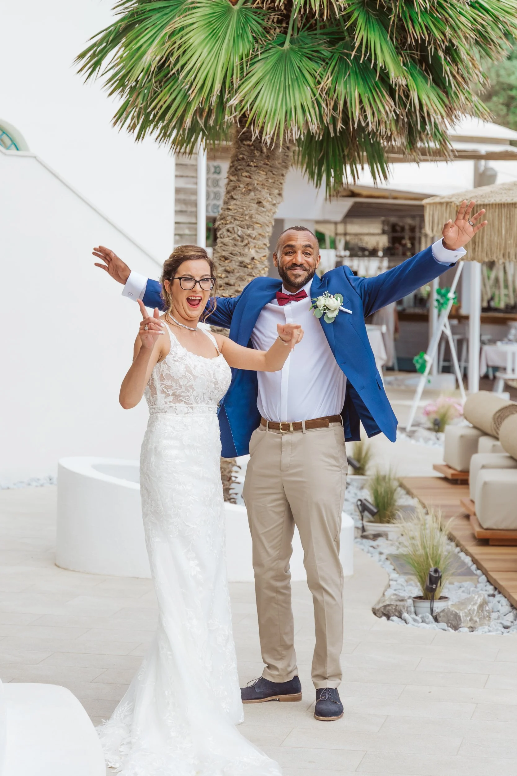 A newlywed couple celebrating at their wedding with the bride in a white lace wedding gown and the groom in a blue suit with a red bow tie, standing outdoors under a palm tree.
