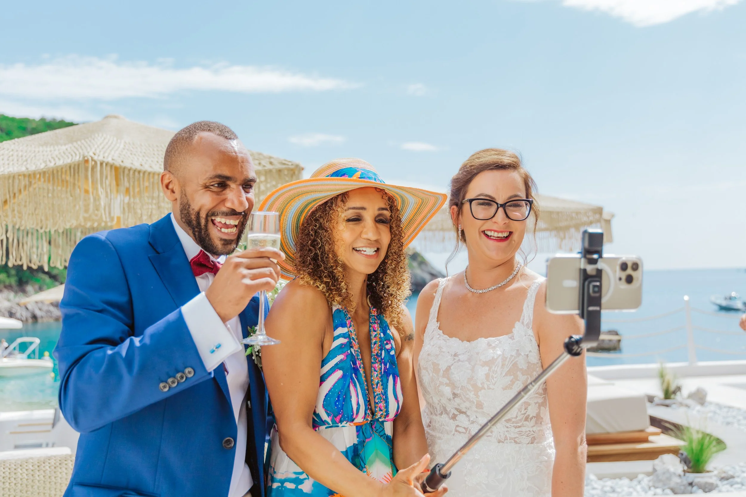 Three friends at a beach party taking a selfie with a smartphone on a selfie stick, smiling and enjoying drinks under beach umbrellas near the ocean.