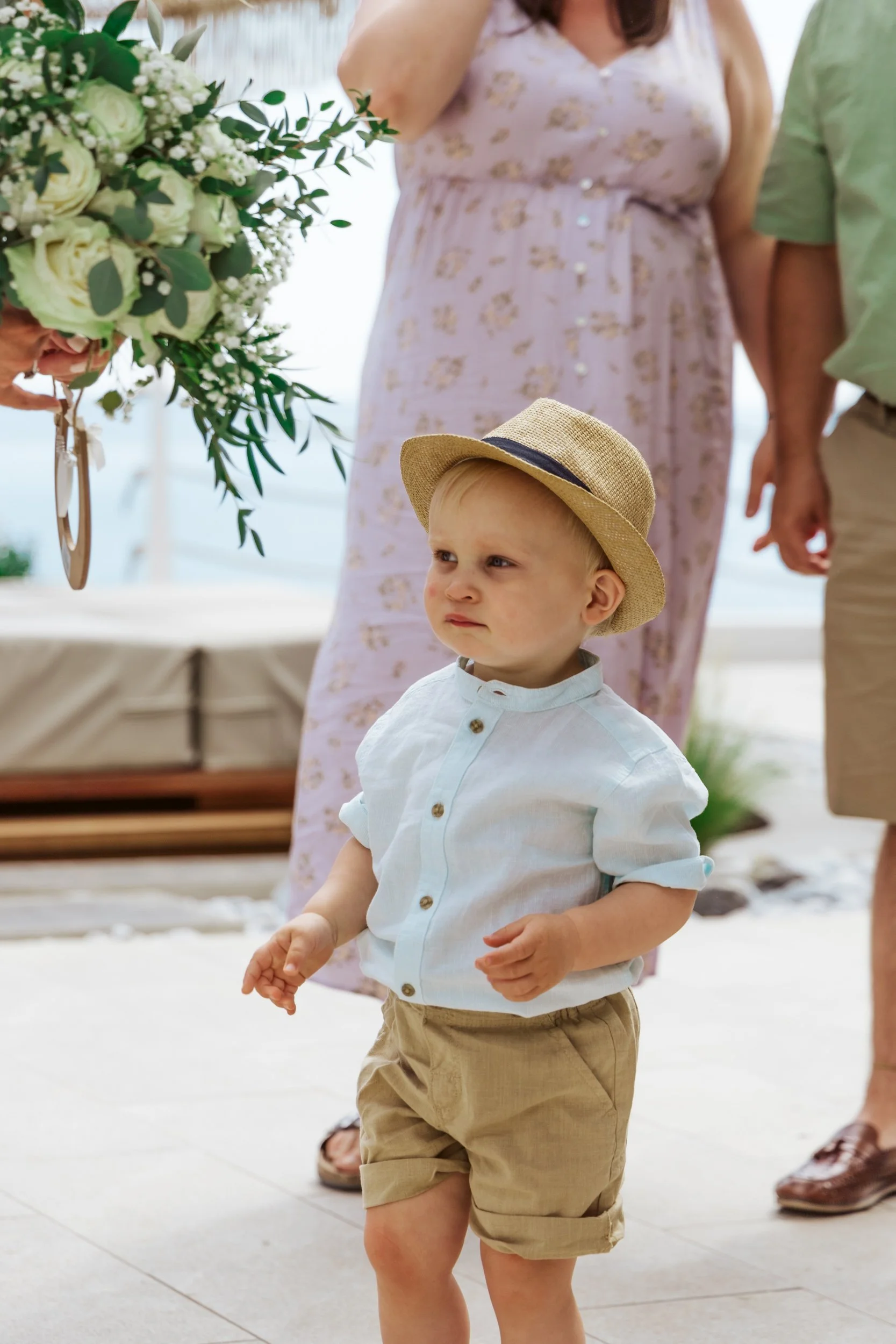A young boy with a serious expression wearing a straw hat, white button-up shirt with rolled sleeves, and tan shorts, standing outdoors near a group of adults, holding a bouquet of flowers.