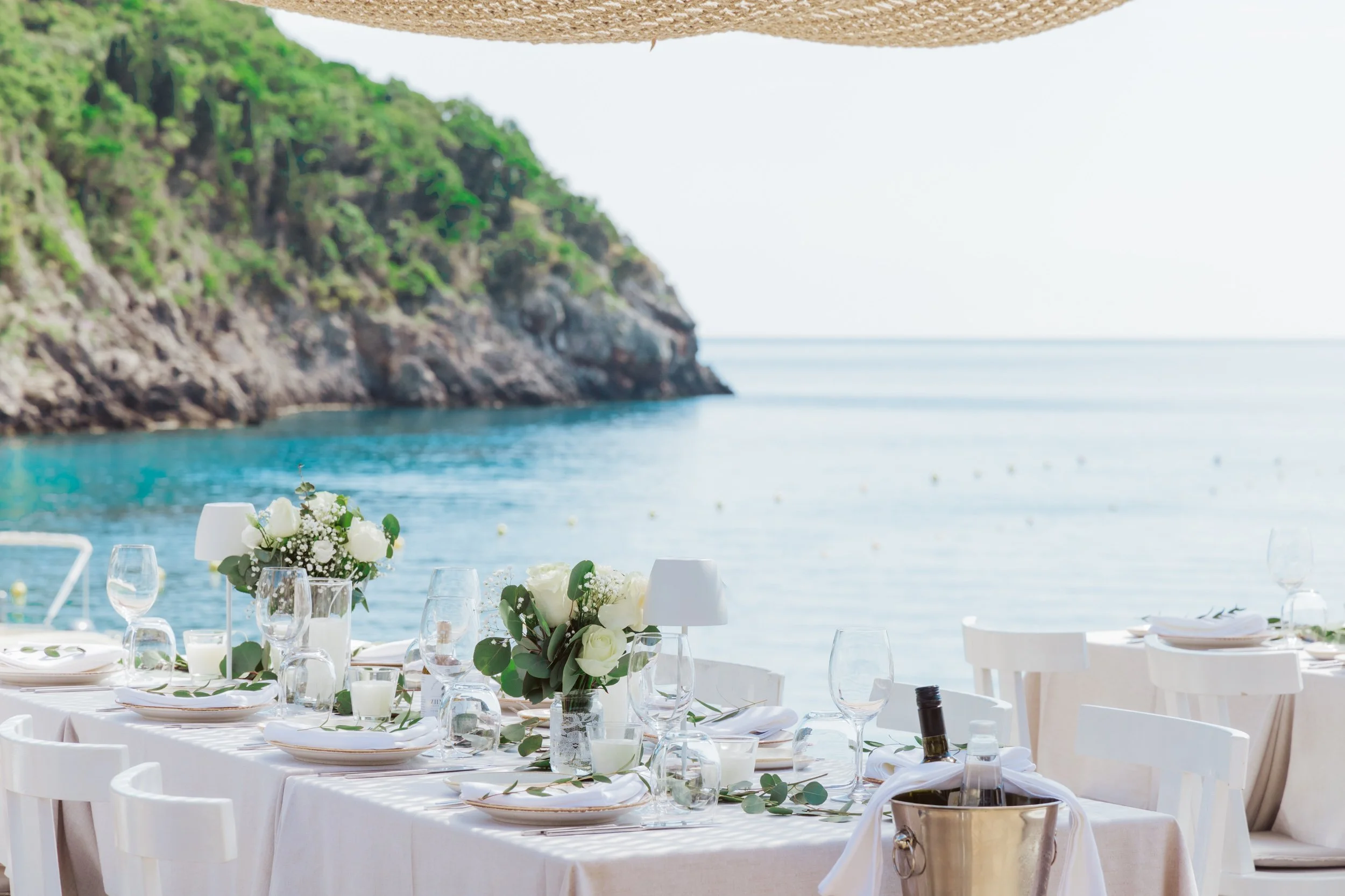 Elegant outdoor dining table set for a meal, decorated with white floral centerpieces, wine glasses, plates, and candles, on a beach with a rocky, green hillside and calm water in the background.