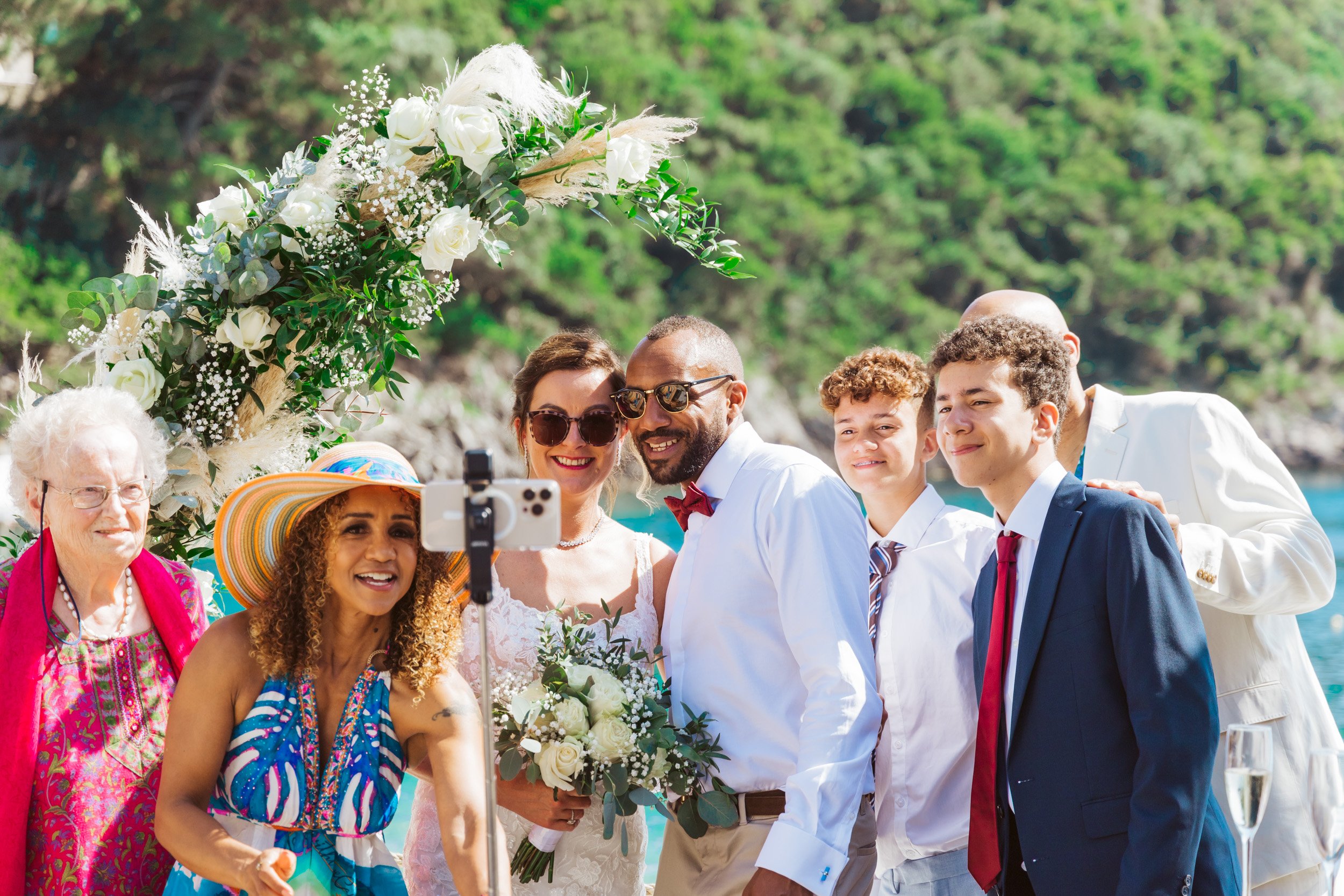 Group of people at a wedding taking a selfie outdoors with a floral arch and greenery background.
