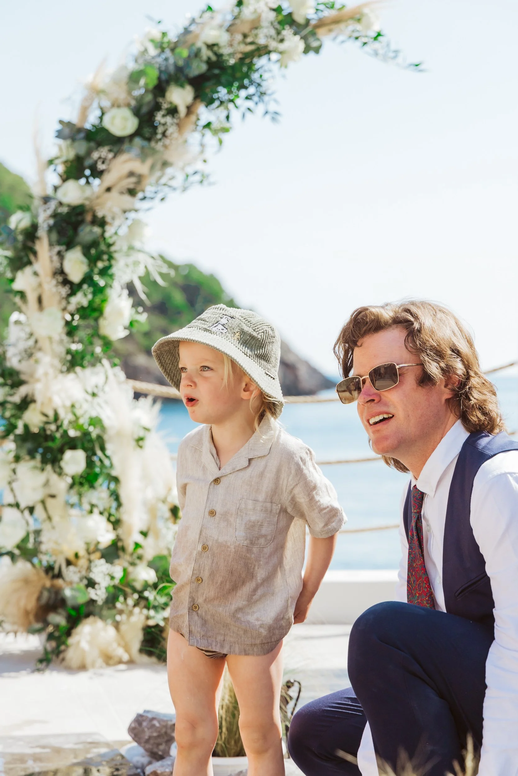 A young boy and man at a beach wedding ceremony; boy wearing a sun hat, beige shirt, and no shorts, standing next to the man with sunglasses, both looking ahead near a floral arch.