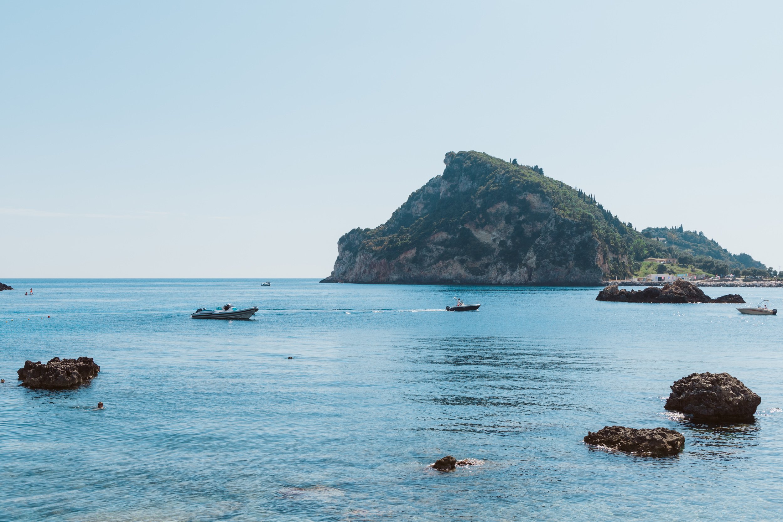 Calm sea with rocks in foreground, several boats floating on water, an island or large hill in the background, clear blue sky.