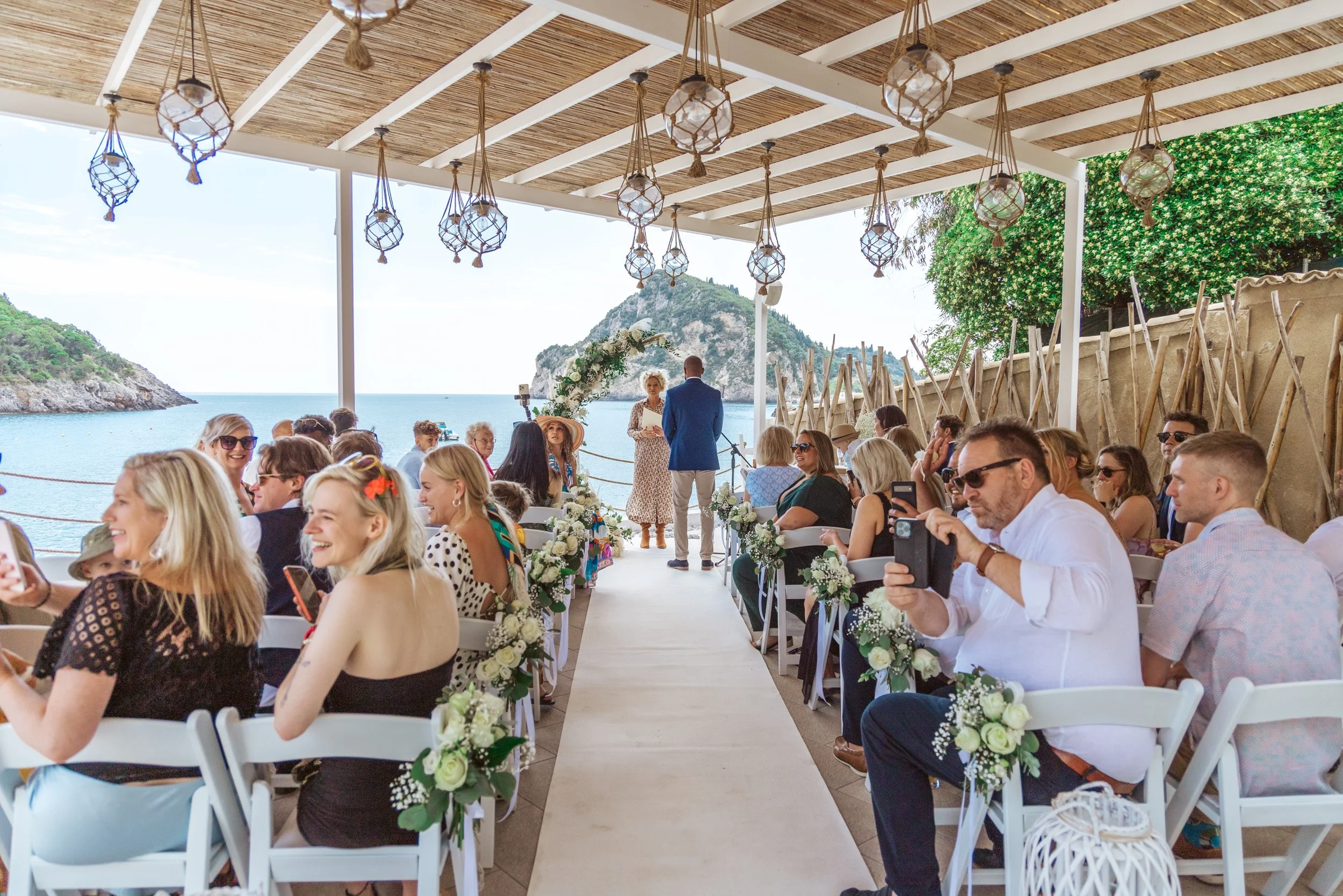 A wedding ceremony taking place at an outdoor beachside venue with guests seated under a wooden roof, overlooking the ocean with mountains in the background. The aisle is decorated with white flowers and greenery.