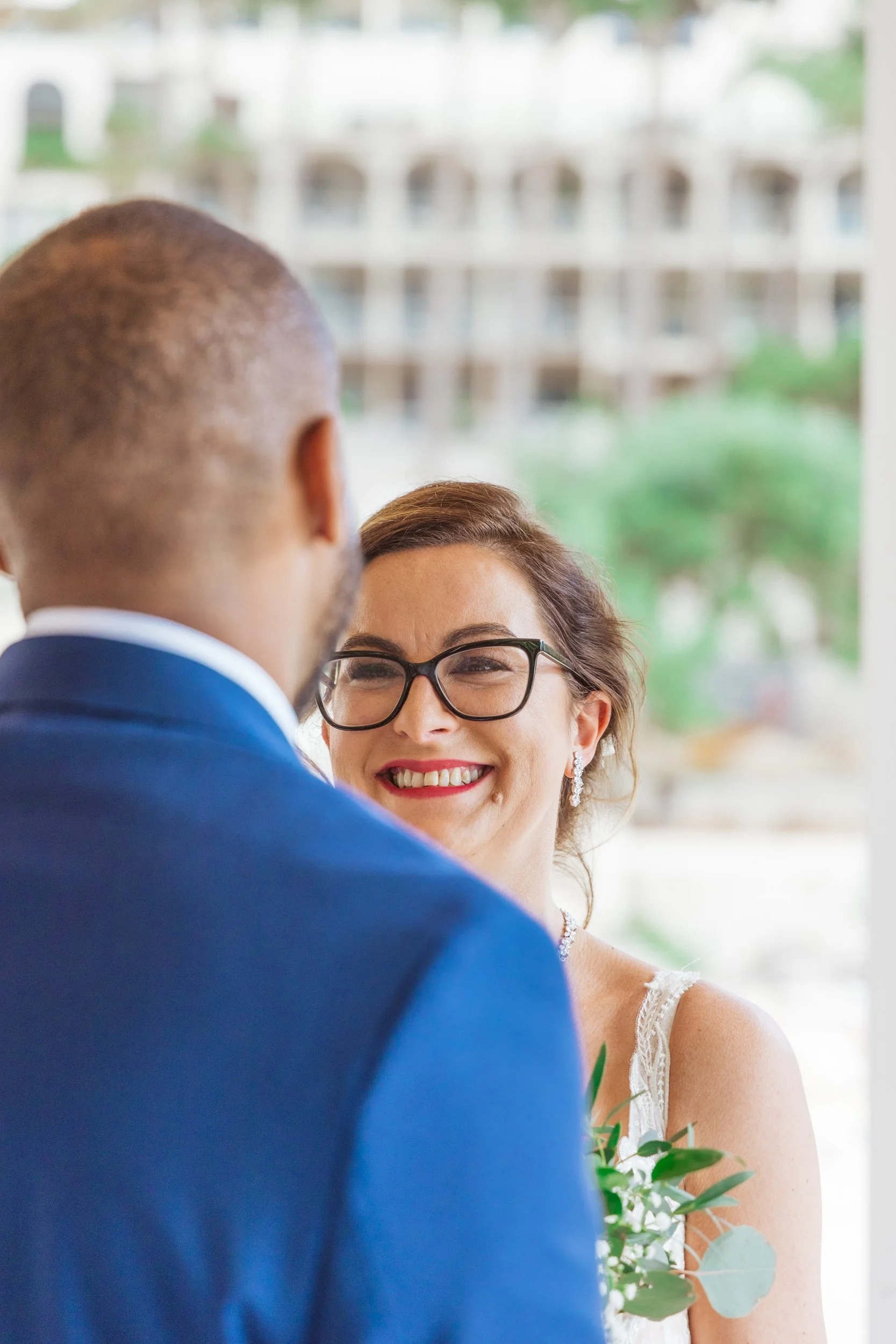 A smiling bride with glasses and earrings looking at her groom during a wedding ceremony, with a blurred cityscape background.