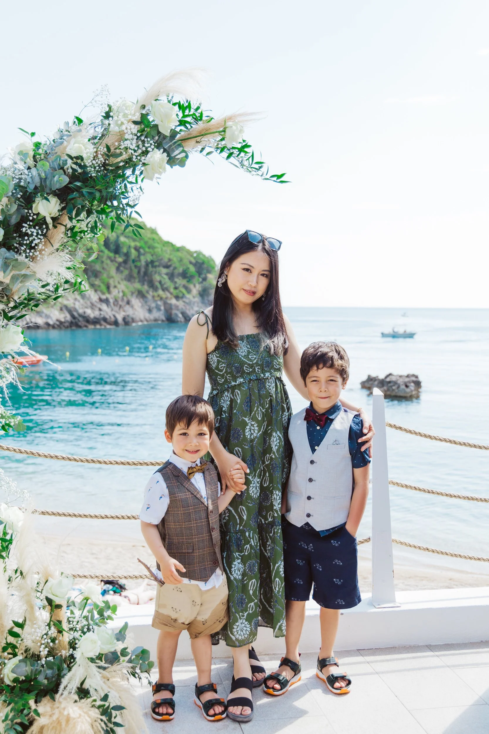A woman and two young boys stand on a beachside promenade with the ocean and a rocky coastline in the background, near an arch decorated with white flowers and greenery.