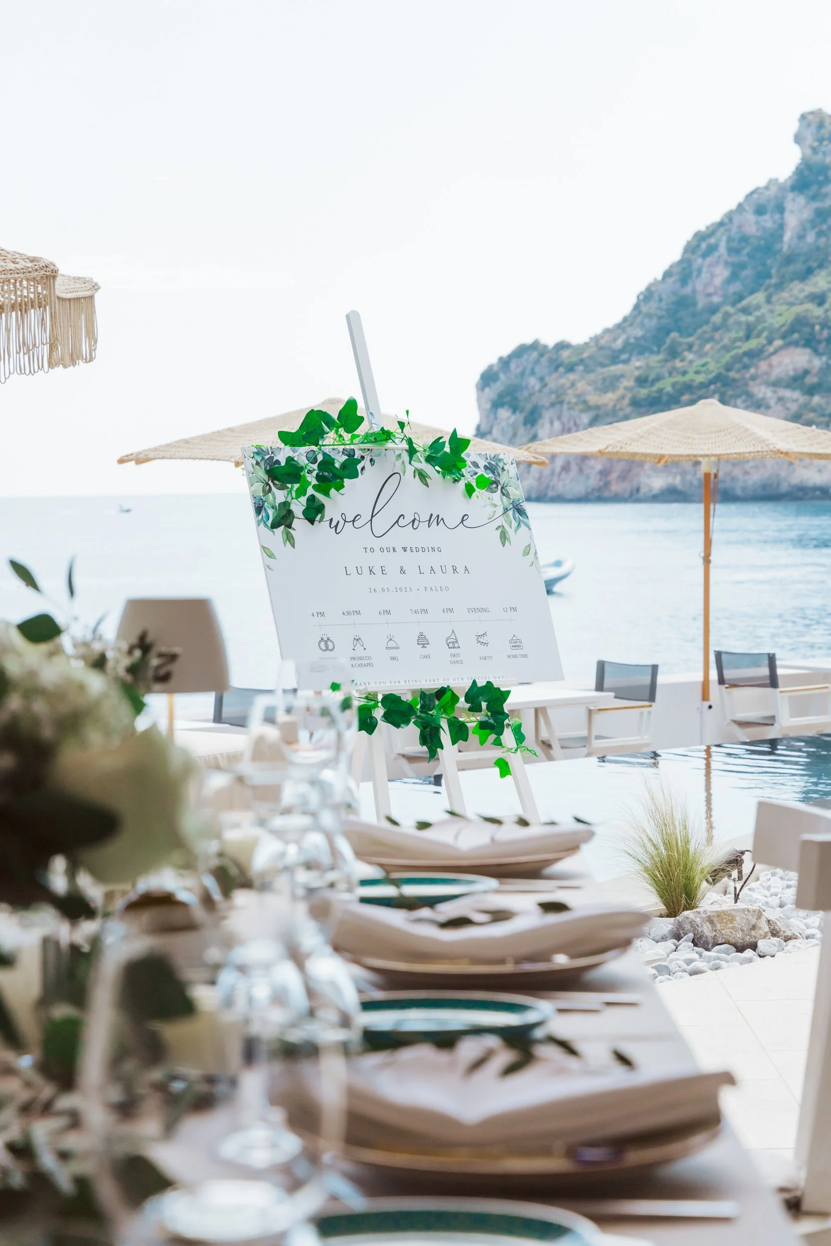 Beachside wedding reception with a table set and a welcome sign for Luke and Laura, overlooking the water with umbrellas and a mountain in the background.