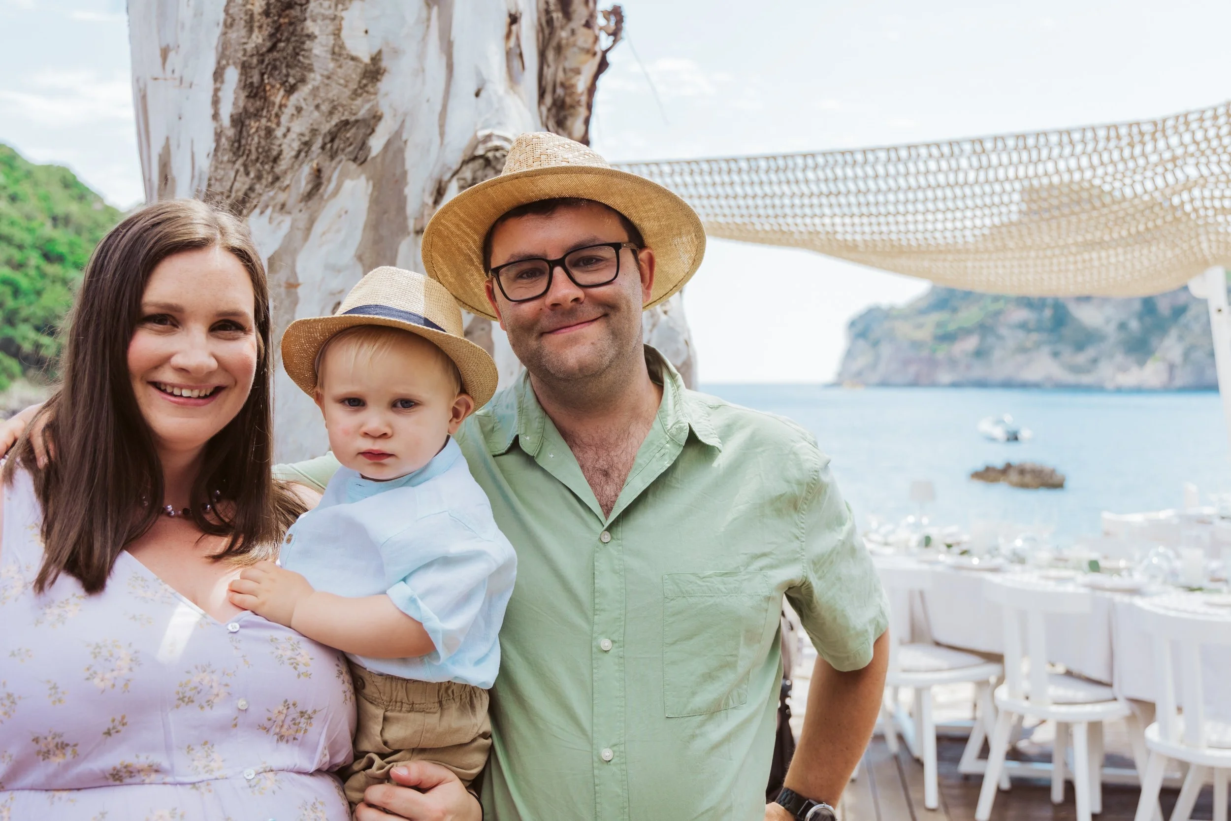 A family of three, a woman, a man, and a young boy, standing close together outdoors near a large tree with a view of the ocean and cliffs in the background. They are dressed in summer clothing and wearing straw hats, smiling for the camera.
