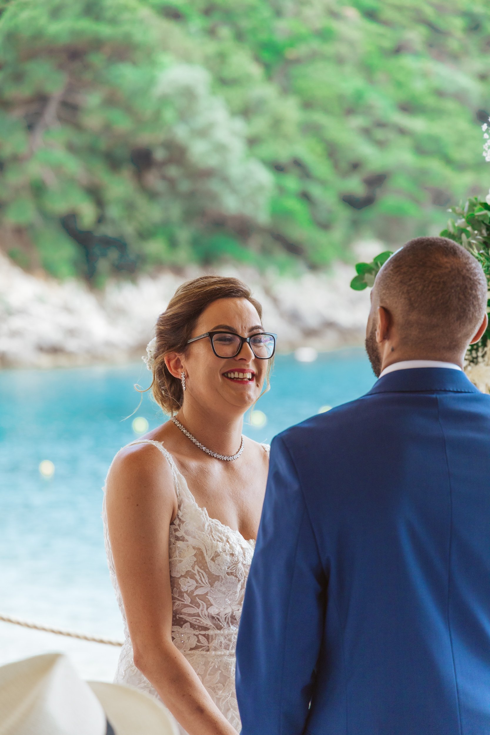 A woman in a white lace wedding dress with glasses and jewelry smiling at a man in a blue suit during a wedding ceremony by a waterfront with green trees in the background.