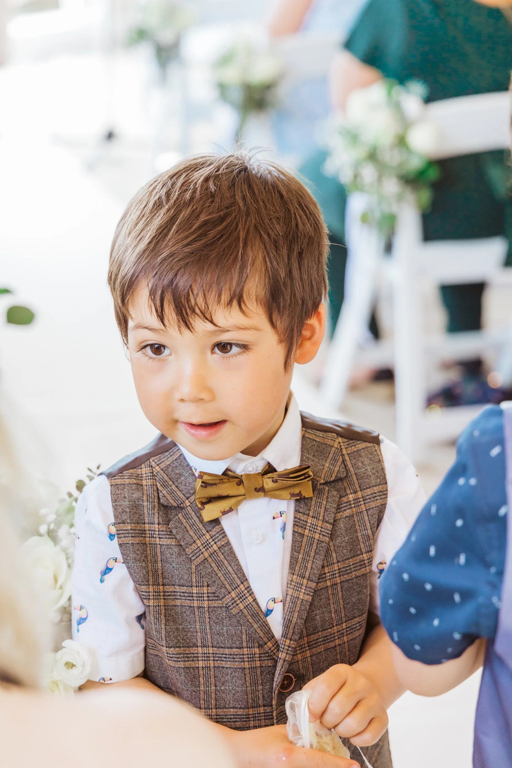 Young boy with brown hair wearing a plaid vest, a white shirt with toucan print, and a yellow bow tie, interacting with another person at a social event.