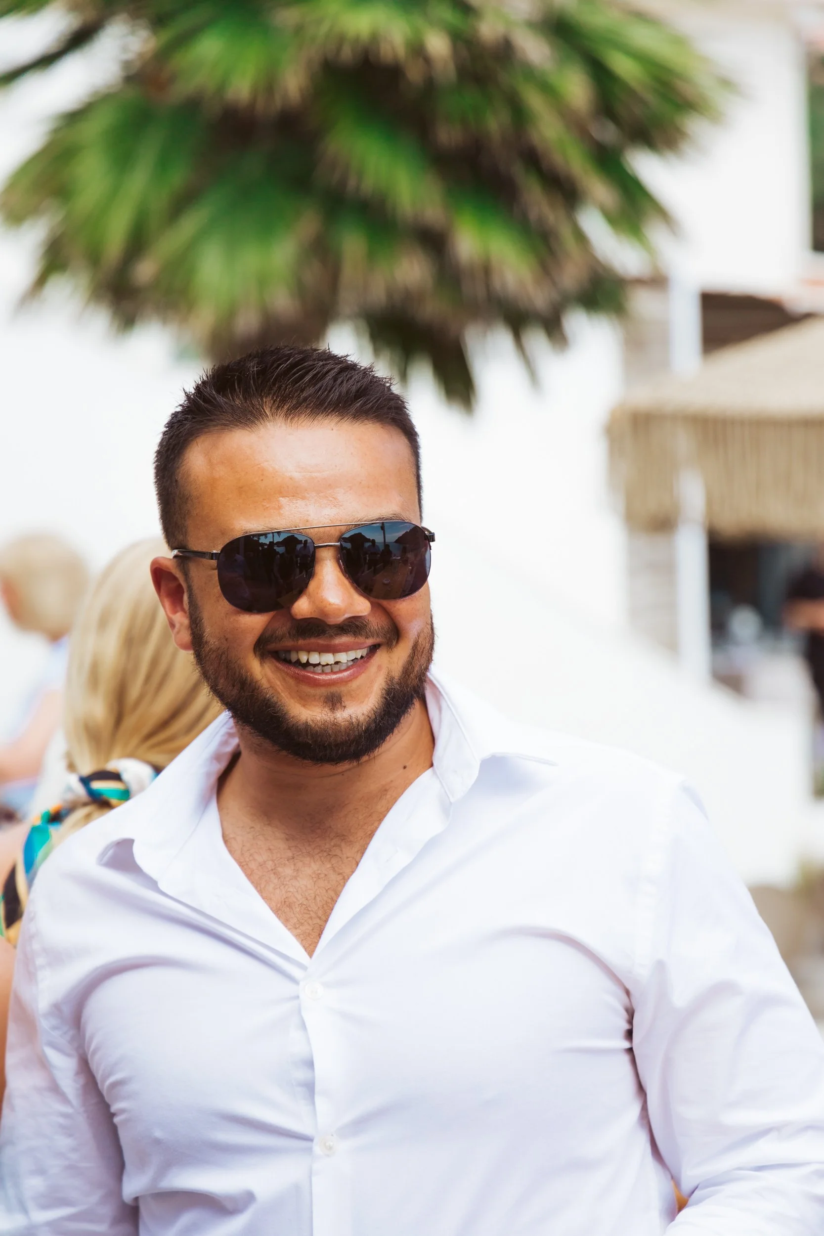 Smiling man with dark hair and beard wearing sunglasses and a white shirt at an outdoor event.