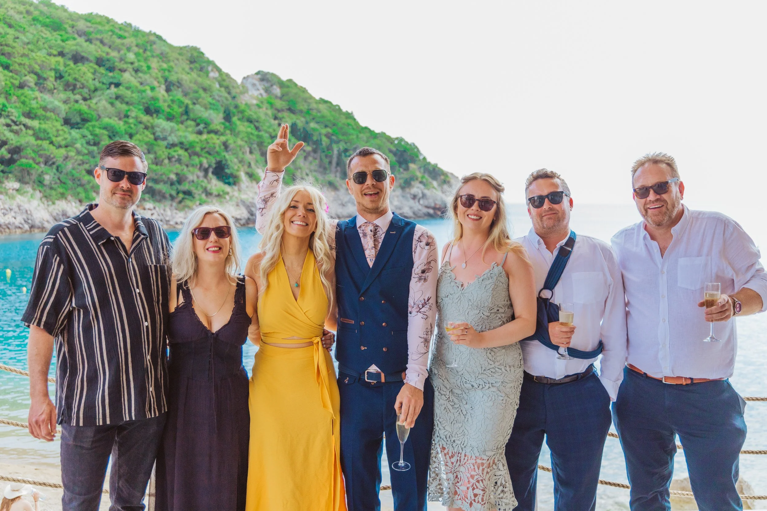 Group of seven people in summer clothes and sunglasses, standing on a boat or near water, with a green hillside and blue ocean in the background, celebrating with drinks.
