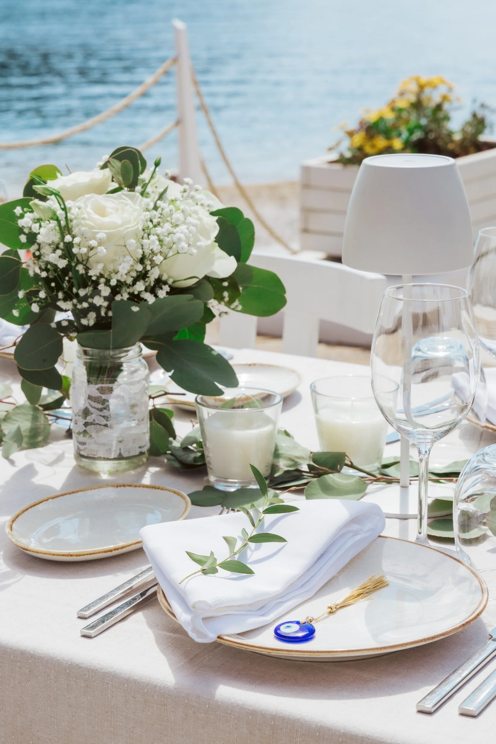 Table set for a meal with a floral centerpiece, candles, glassware, plates, and white napkins, overlooking the sea with a white wooden railing and potted plants in the background.