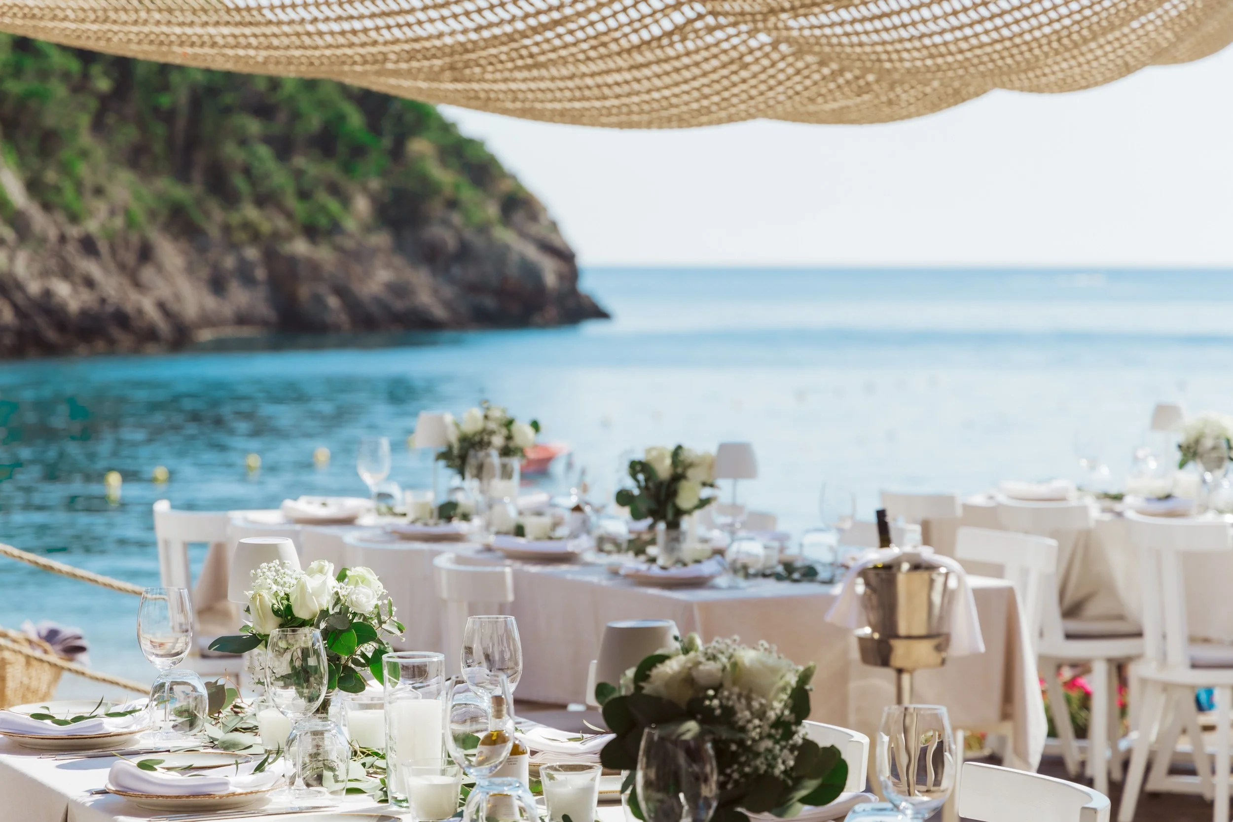 Elegant outdoor seaside dining setup with white tables, chairs, and floral centerpieces, overlooking the ocean with a rocky cliff in the background.