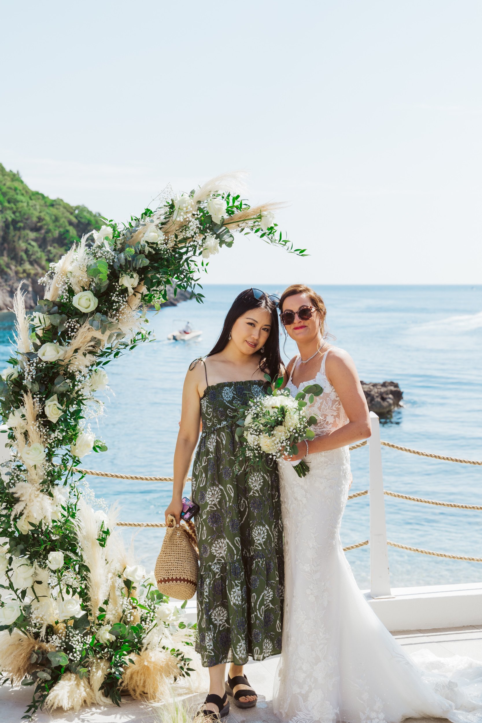Two women, one in a white lace wedding dress holding a bouquet and the other in a green patterned dress holding a woven bag and phone, standing together by the ocean under a floral arch with greenery and white flowers, on a sunny day.