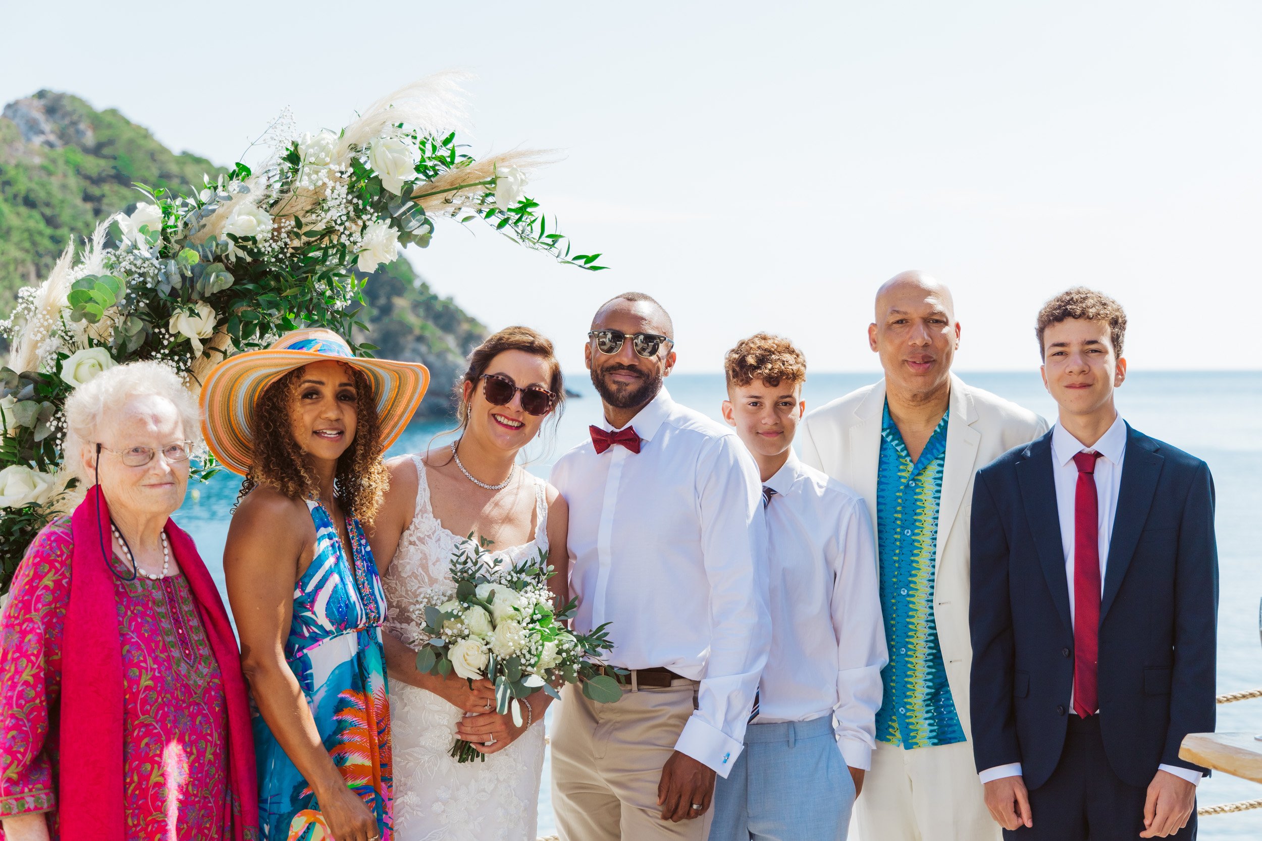 Group of people at a wedding on the beach, including a bride holding a bouquet, surrounded by family and friends near a floral arch with the ocean and mountainous landscape in the background.