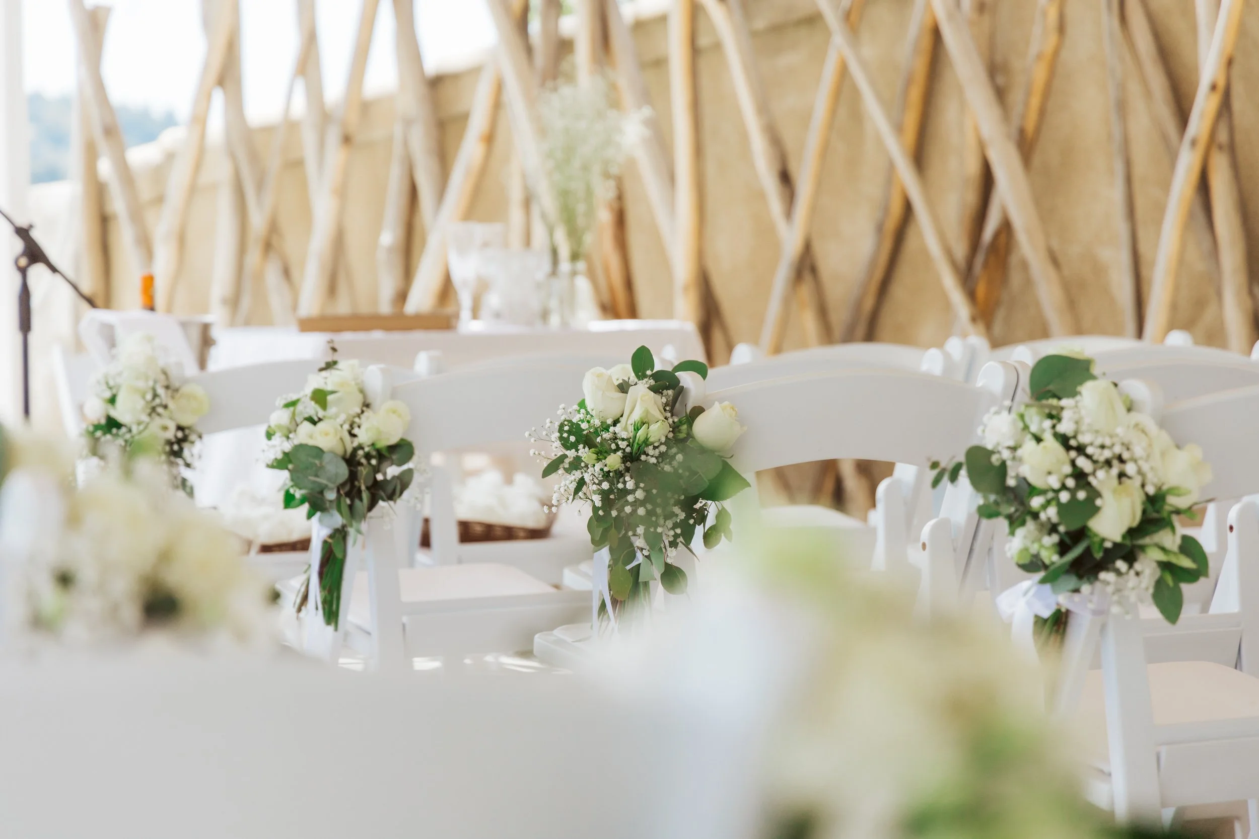 White chairs decorated with small bouquets of white roses and greenery, set up for a wedding or event, with a rustic wooden backdrop.