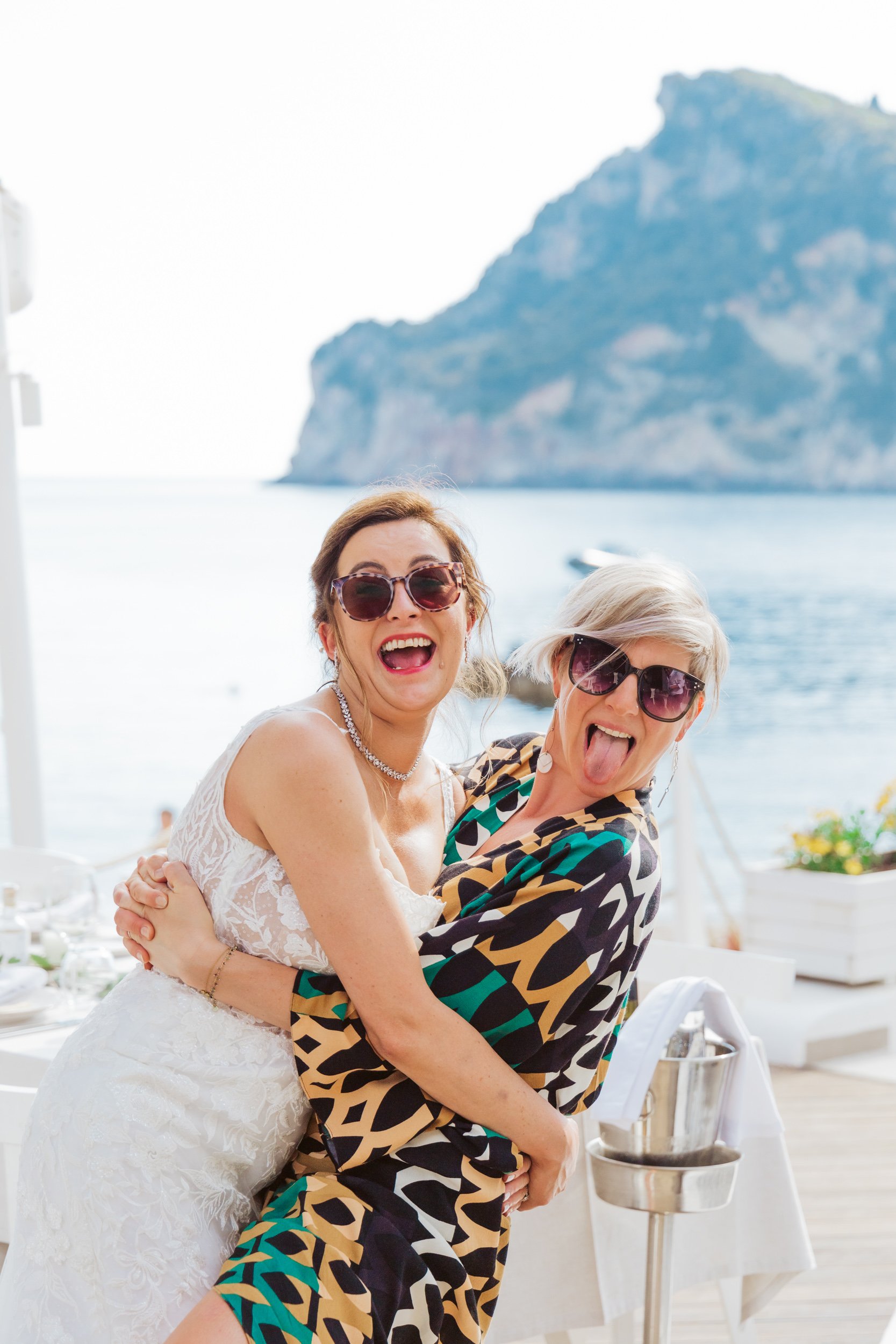 Two women wearing sunglasses having fun, one in a wedding dress and the other in a colorful dress, at an outdoor event with water and mountain in the background.