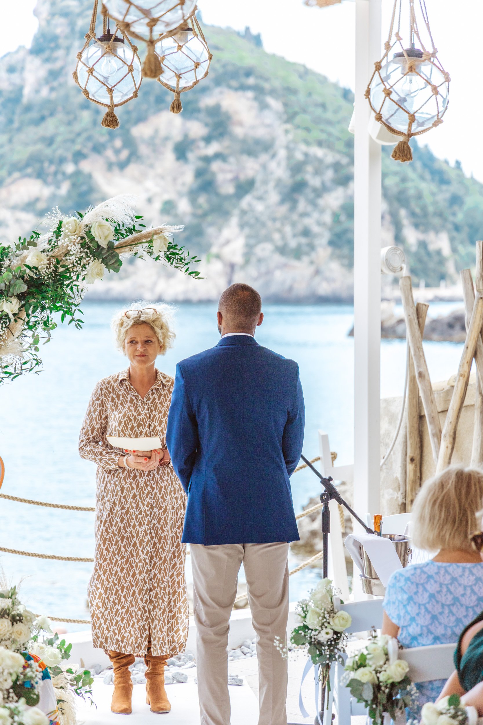 A wedding ceremony taking place outdoors near water with mountains in the background, featuring a woman officiant holding a book and a man dressed in a blue blazer facing her, with guests seated nearby.