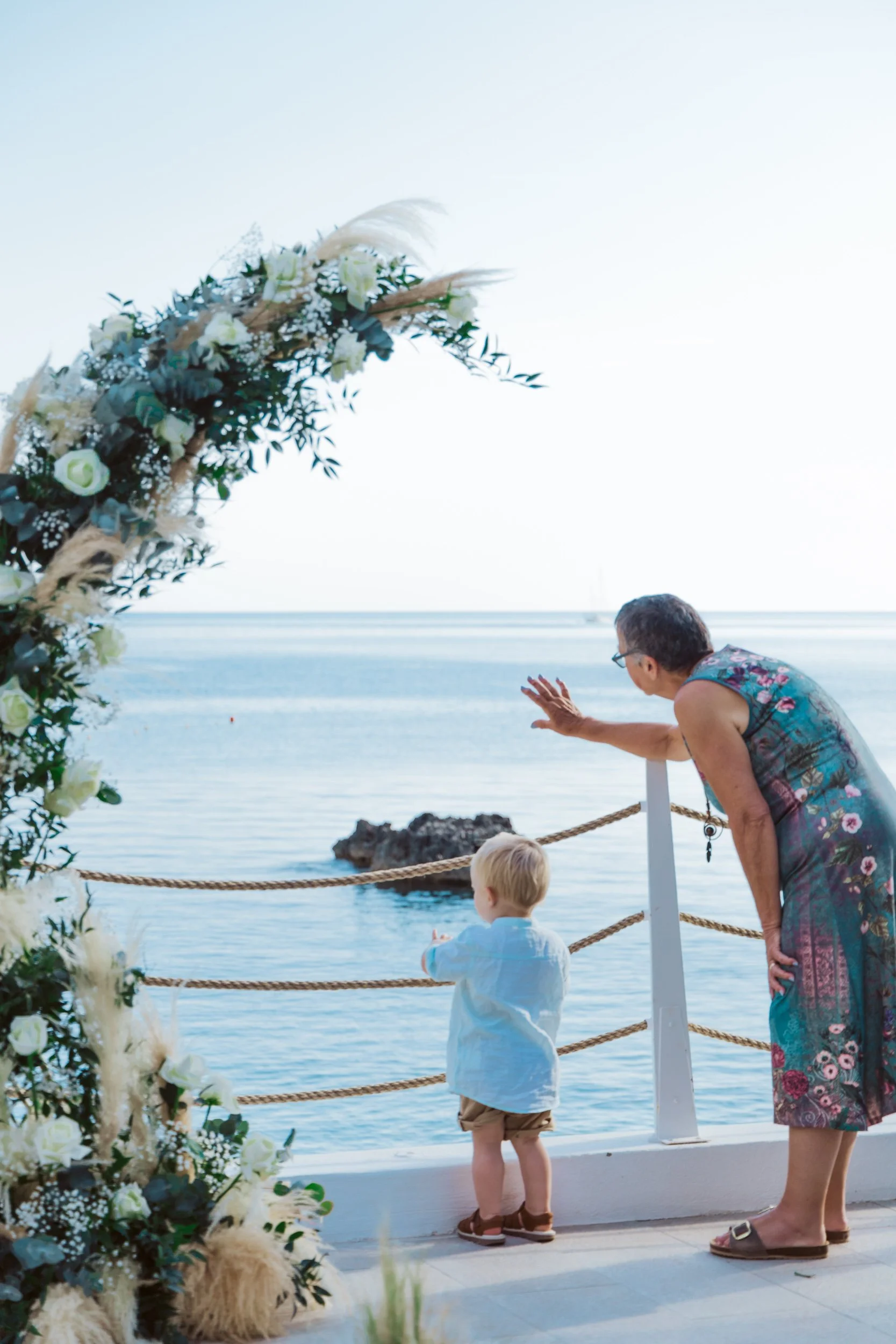 An elderly woman and a young boy looking at the ocean from a decorated outdoor wedding venue with floral decorations and an arch.