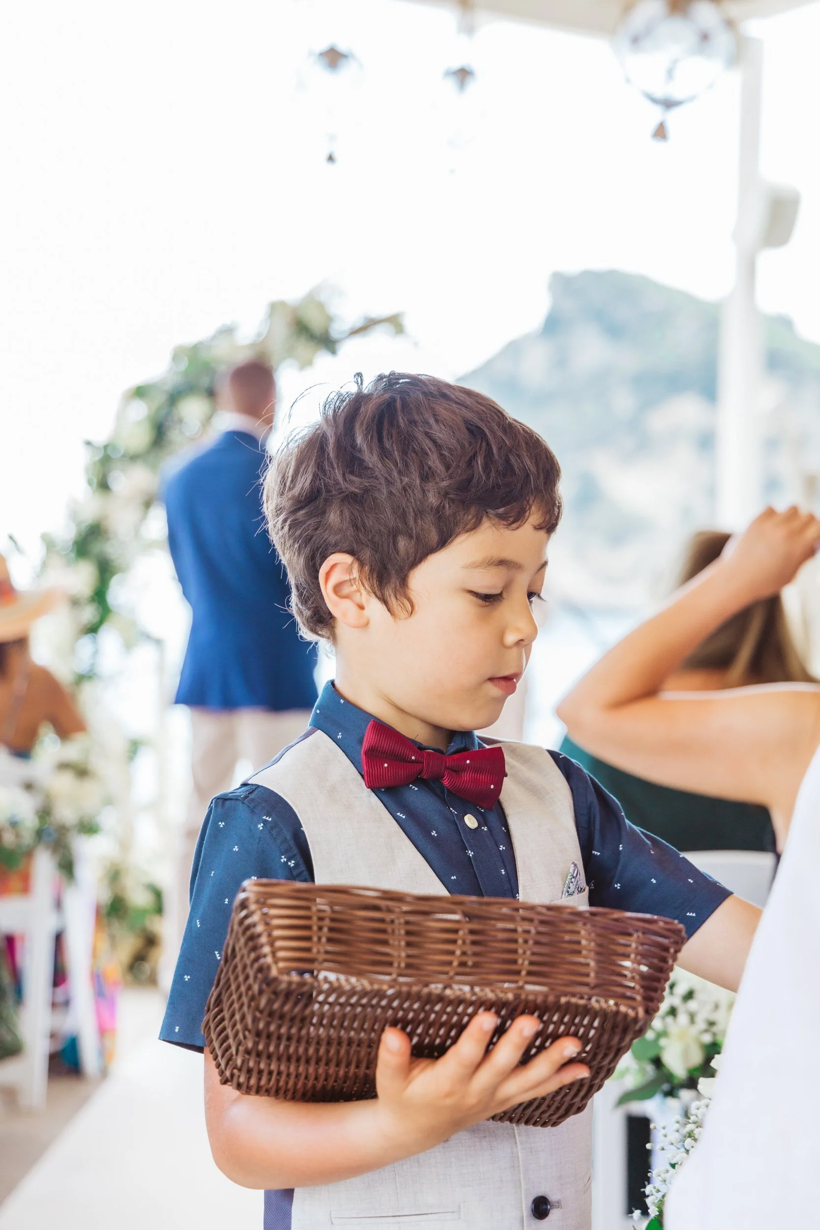 A young boy dressed in a blue shirt with a red bow tie and gray vest, holding a wicker basket, at a wedding reception with blurred guests and decorations in the background.