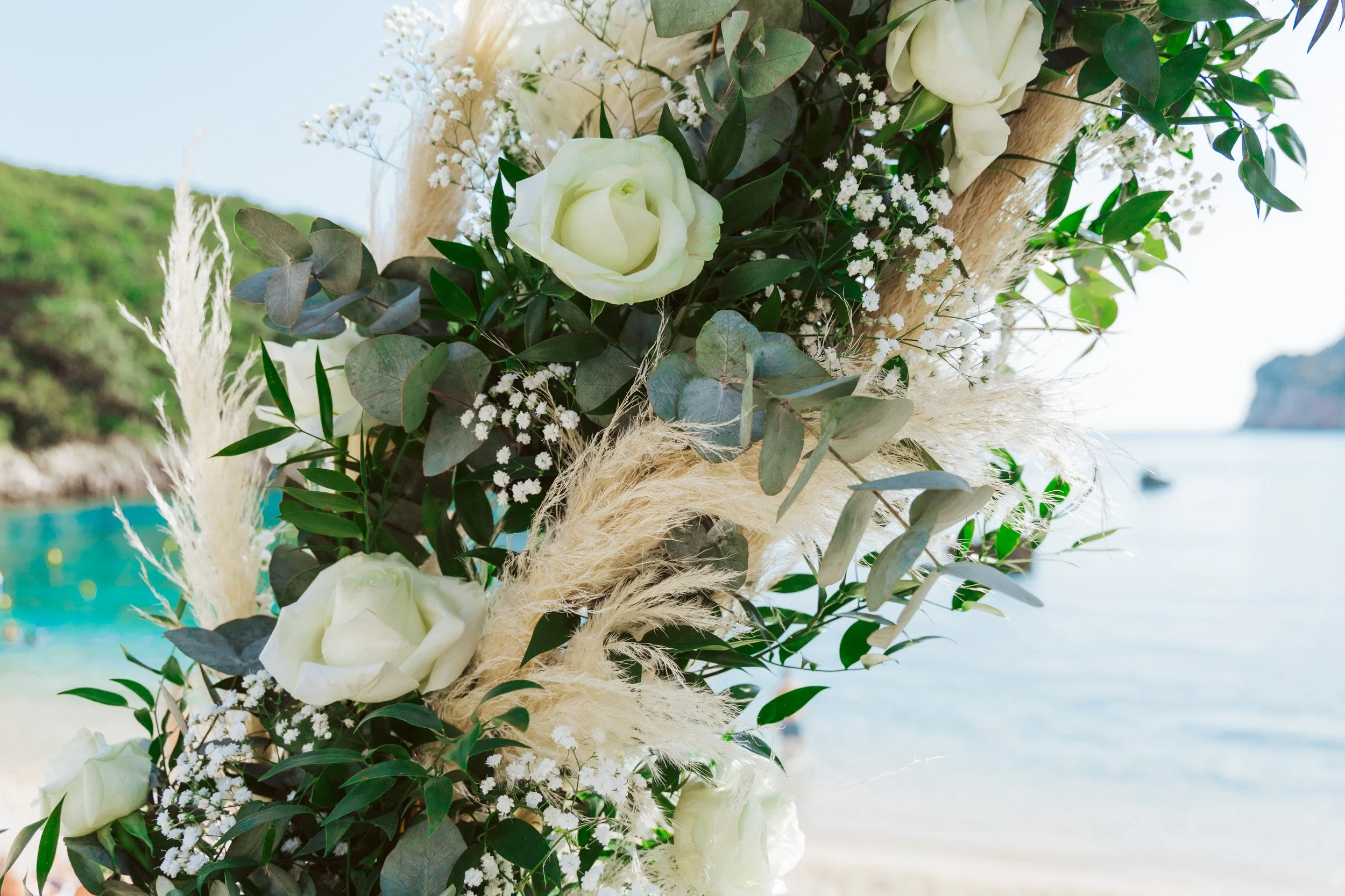 Close-up of a floral arrangement with white roses, eucalyptus leaves, baby's breath, and pampas grass on a beach with water and green hills in the background.