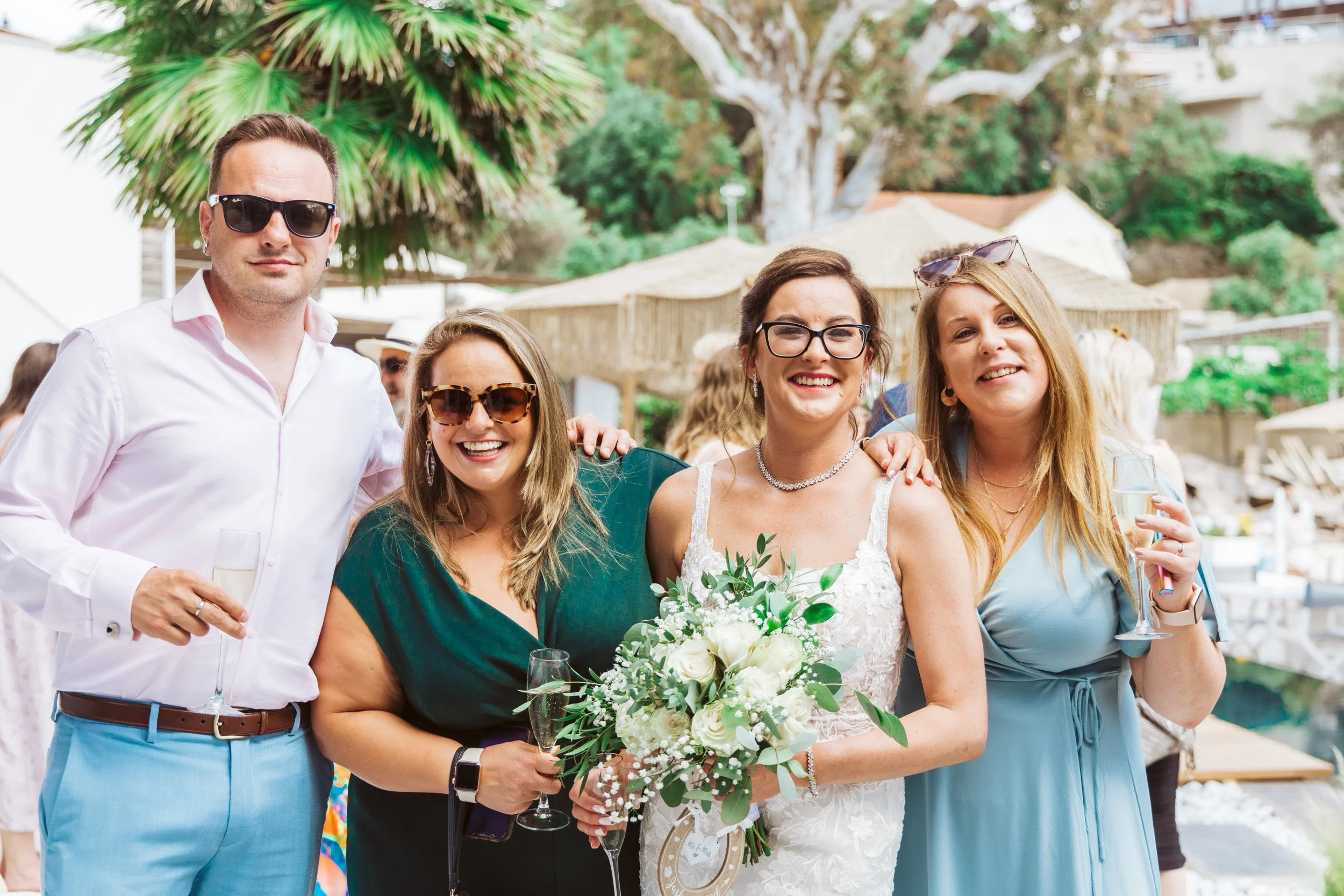 Group of five people smiling, at a wedding celebration outdoors, with a bride holding a bouquet of white flowers, and two women holding glasses of champagne.